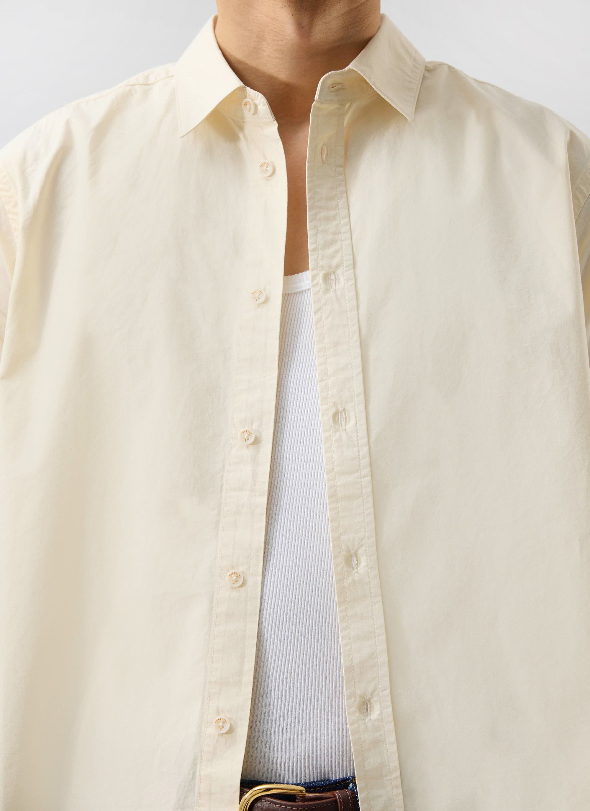 Close up of a model wearing the Percival Boxy Cropped Clerk Shirt in ecru, styled open over a white vest.