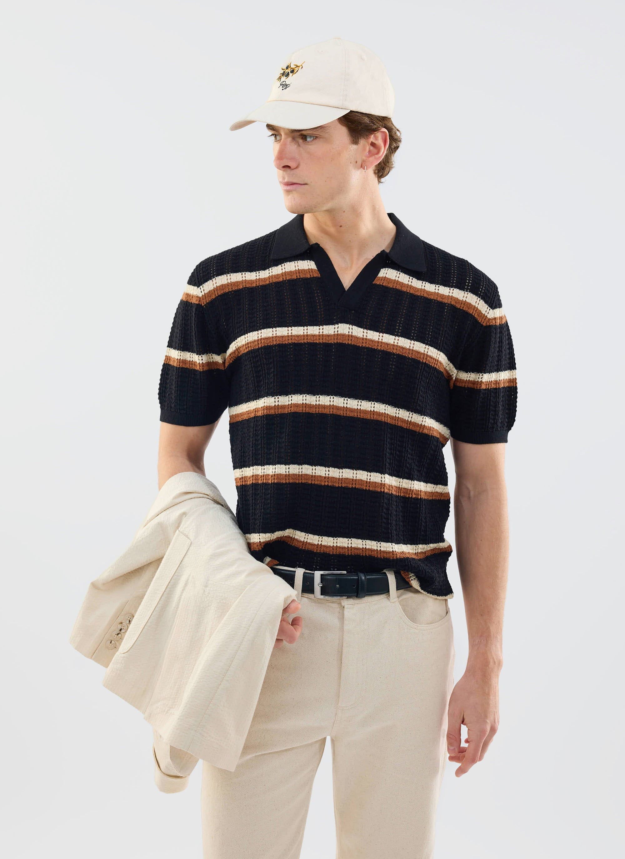 A model wears the black Percival Tessaro Knitted Polo with brown and cream stripes, styled with cream trousers and a cap.