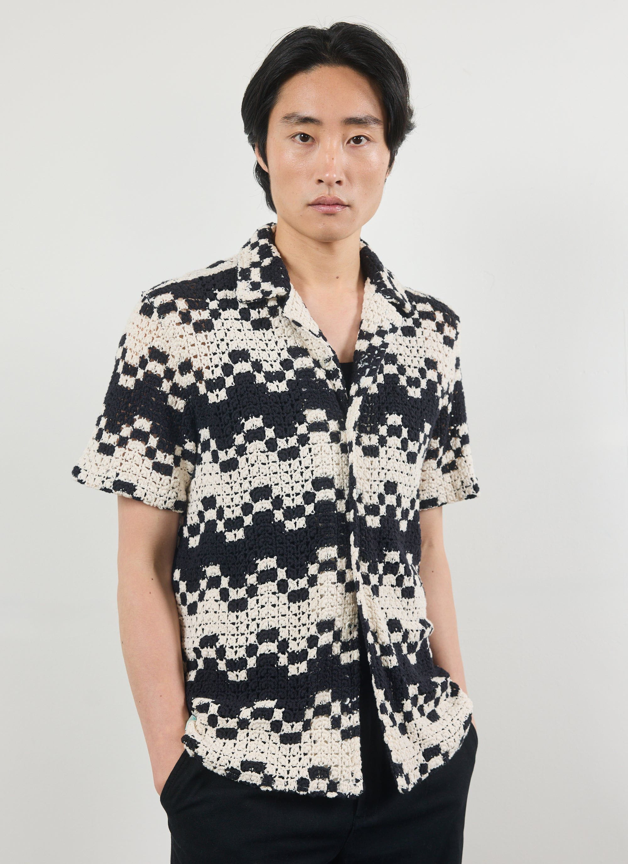 A man models the Percival Naples Crochet Cuban Shirt in an ecru and black geometric crochet knit against a white background.