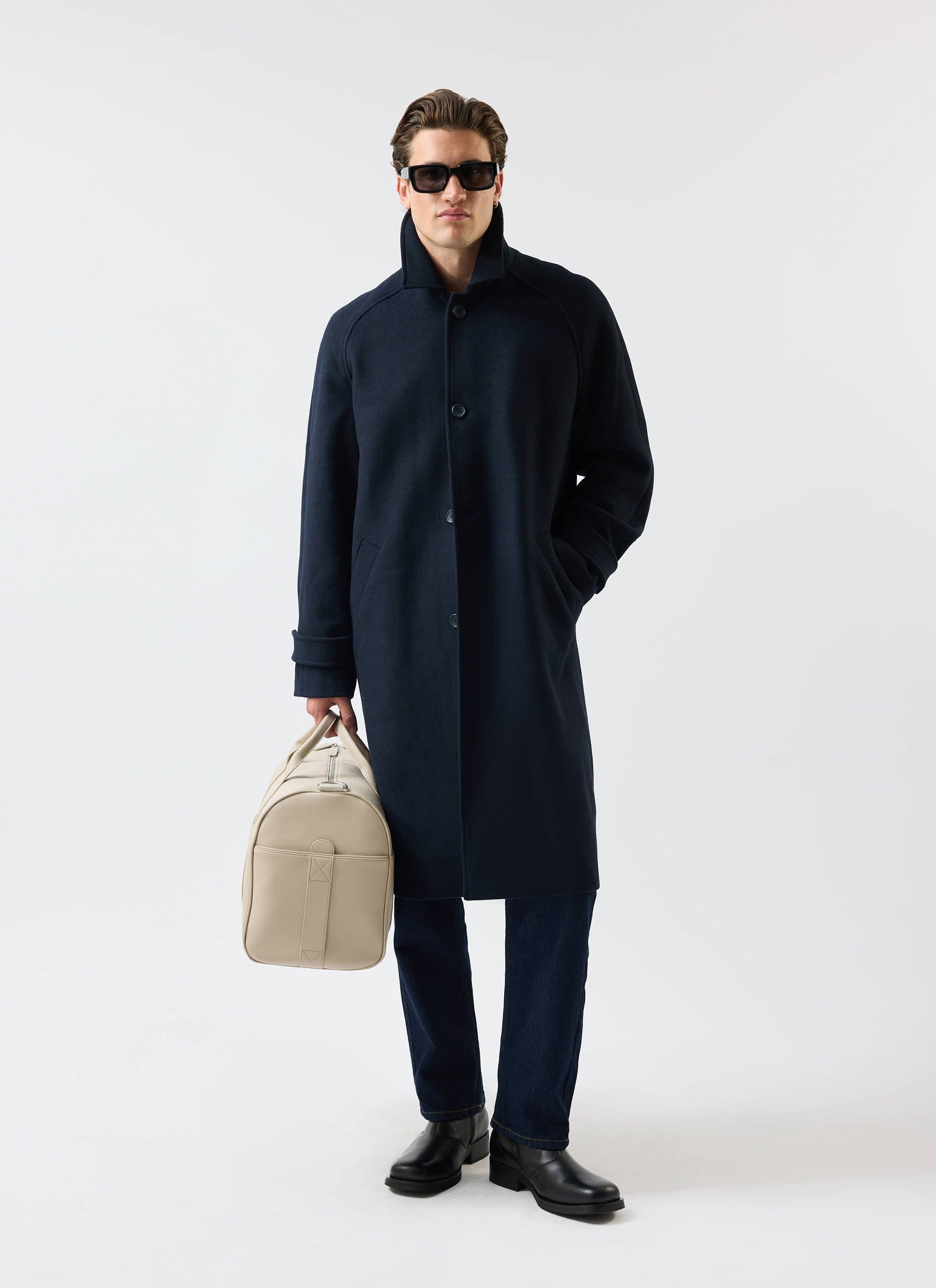 A man models the Percival navy wool Car Coat against a white background, wearing sunglasses and holding a cream weekend bag.