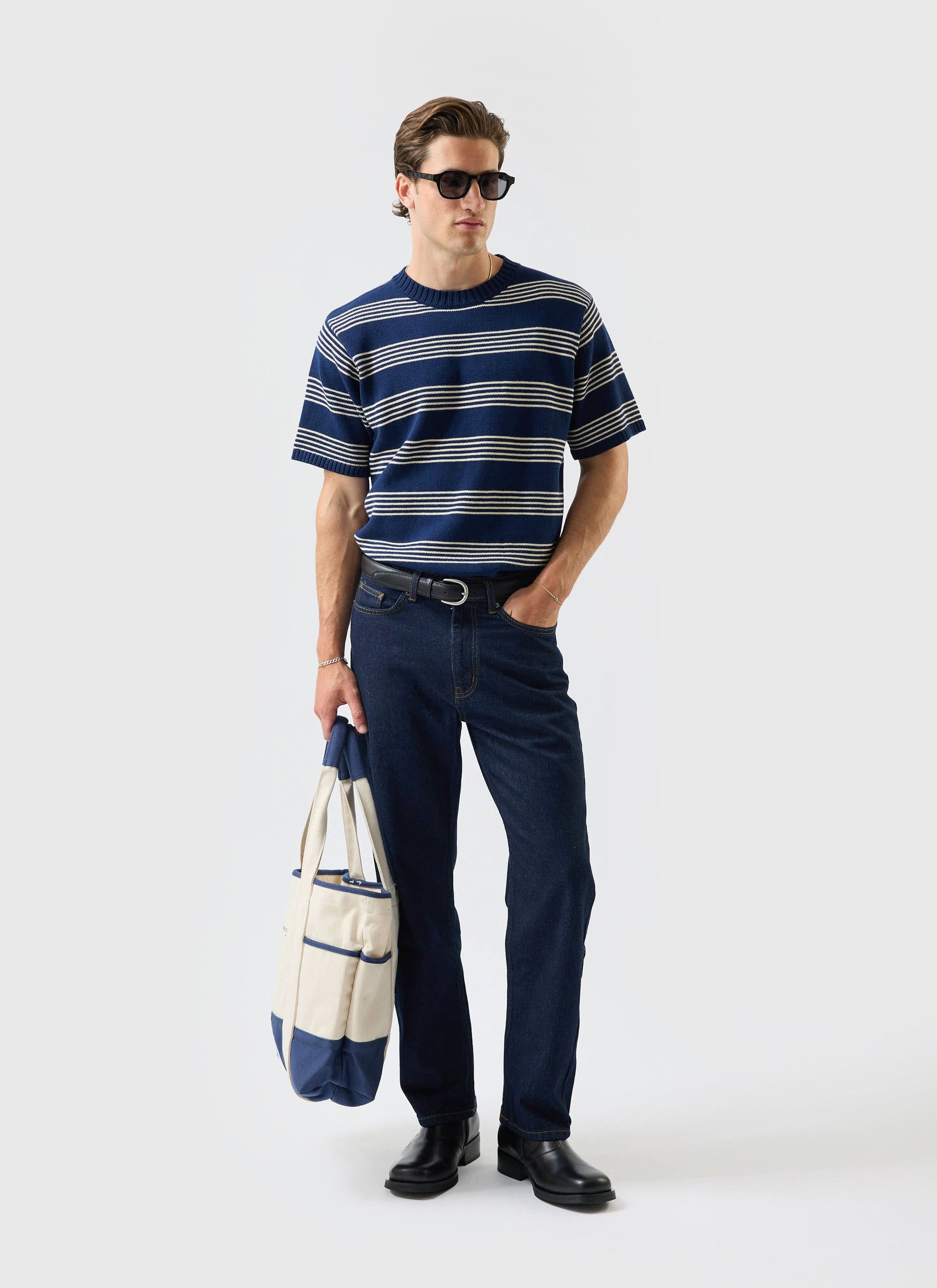 A model wears the Percival Fiction Striped Knitted T Shirt in navy, styled with dark denim jeans, sunglasses and a tote bag.