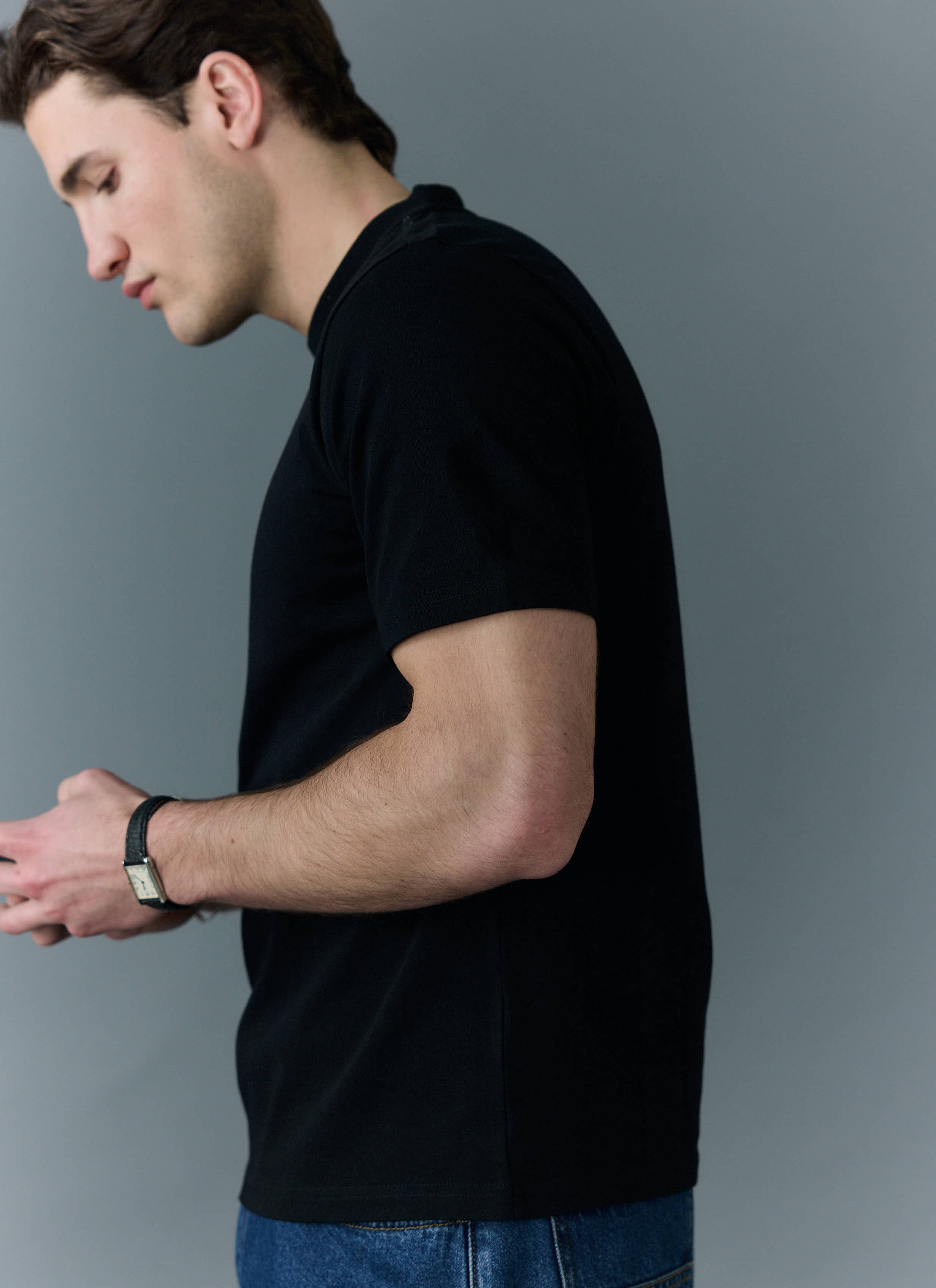 A model wears the Percival Cropped Regular Fit T-shirt in black organic cotton, viewed from the side against a grey wall.