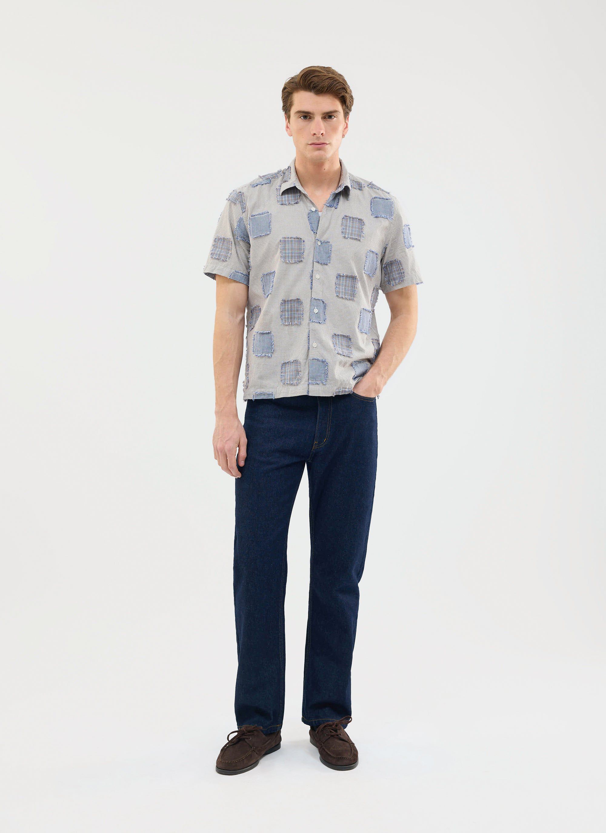 A man models the Percival Bruno Boxy Clerk Shirt in grey, featuring a patchwork design, styled with dark wash jeans.