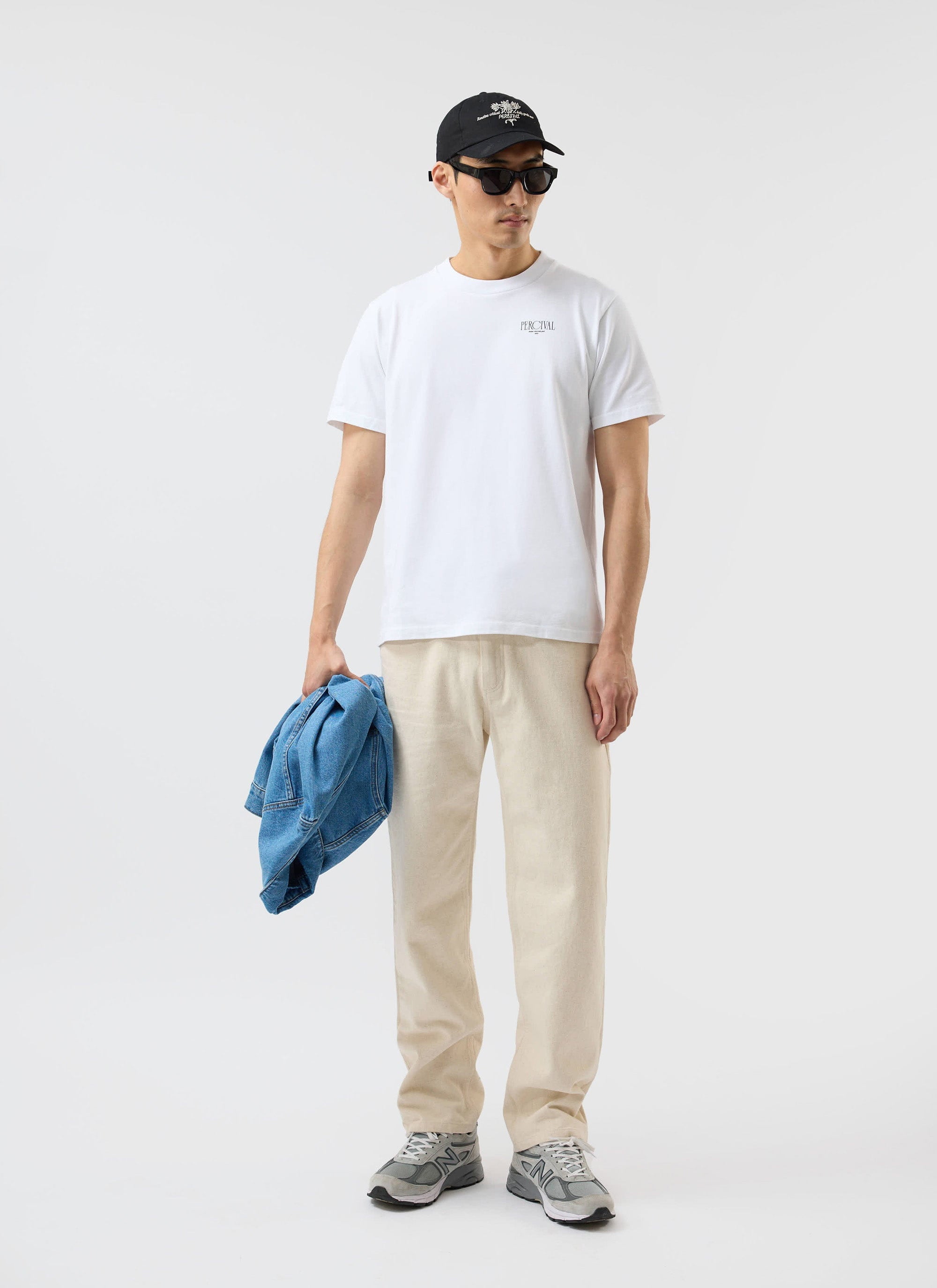 A model wears the white Percival Bistro T Shirt, styled with cream trousers, sunglasses and cap against a white background.