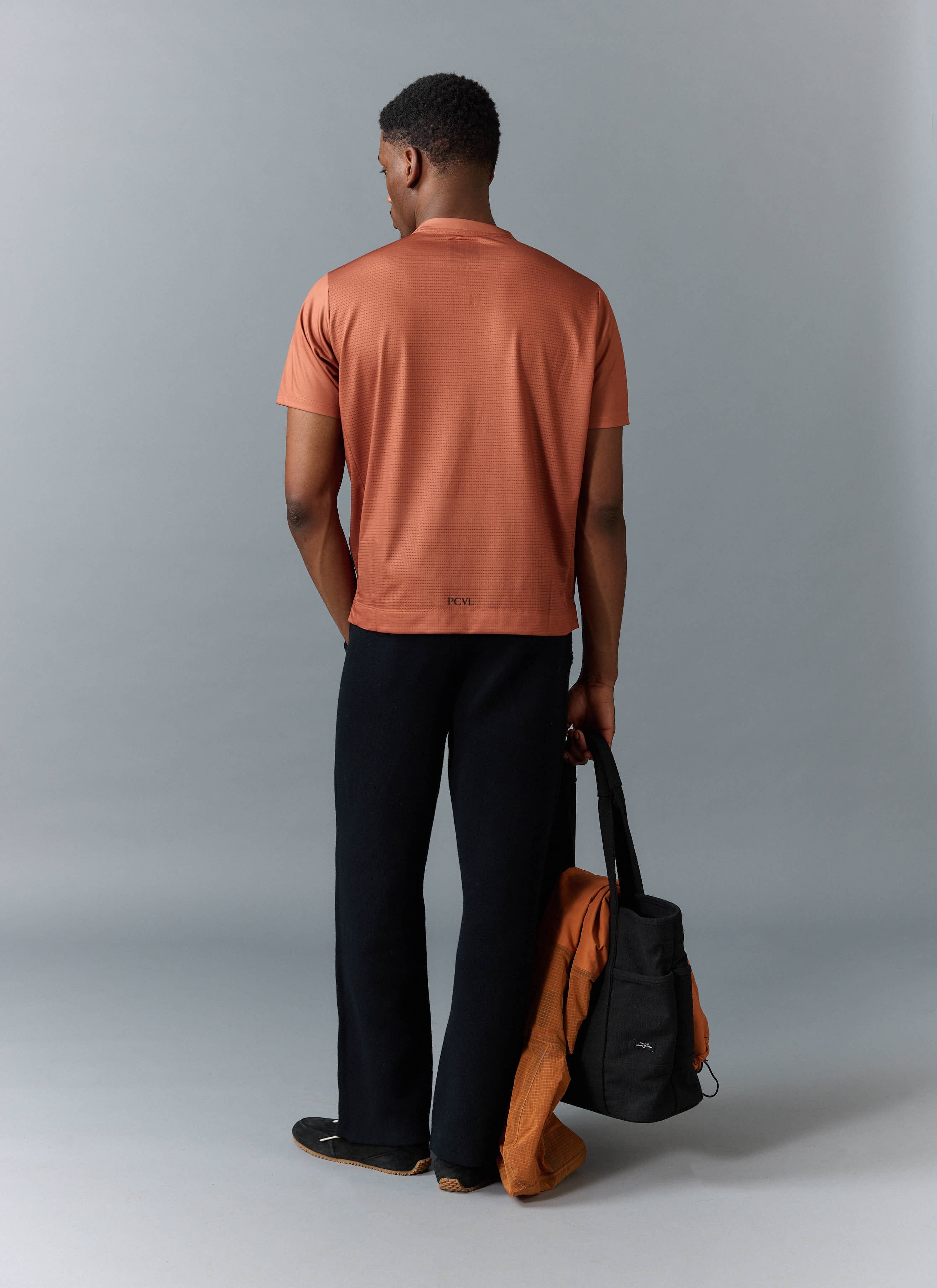 A model seen from behind wears the Percival PCVL Performance Top in rust, styled with black trousers and holding a tote bag.