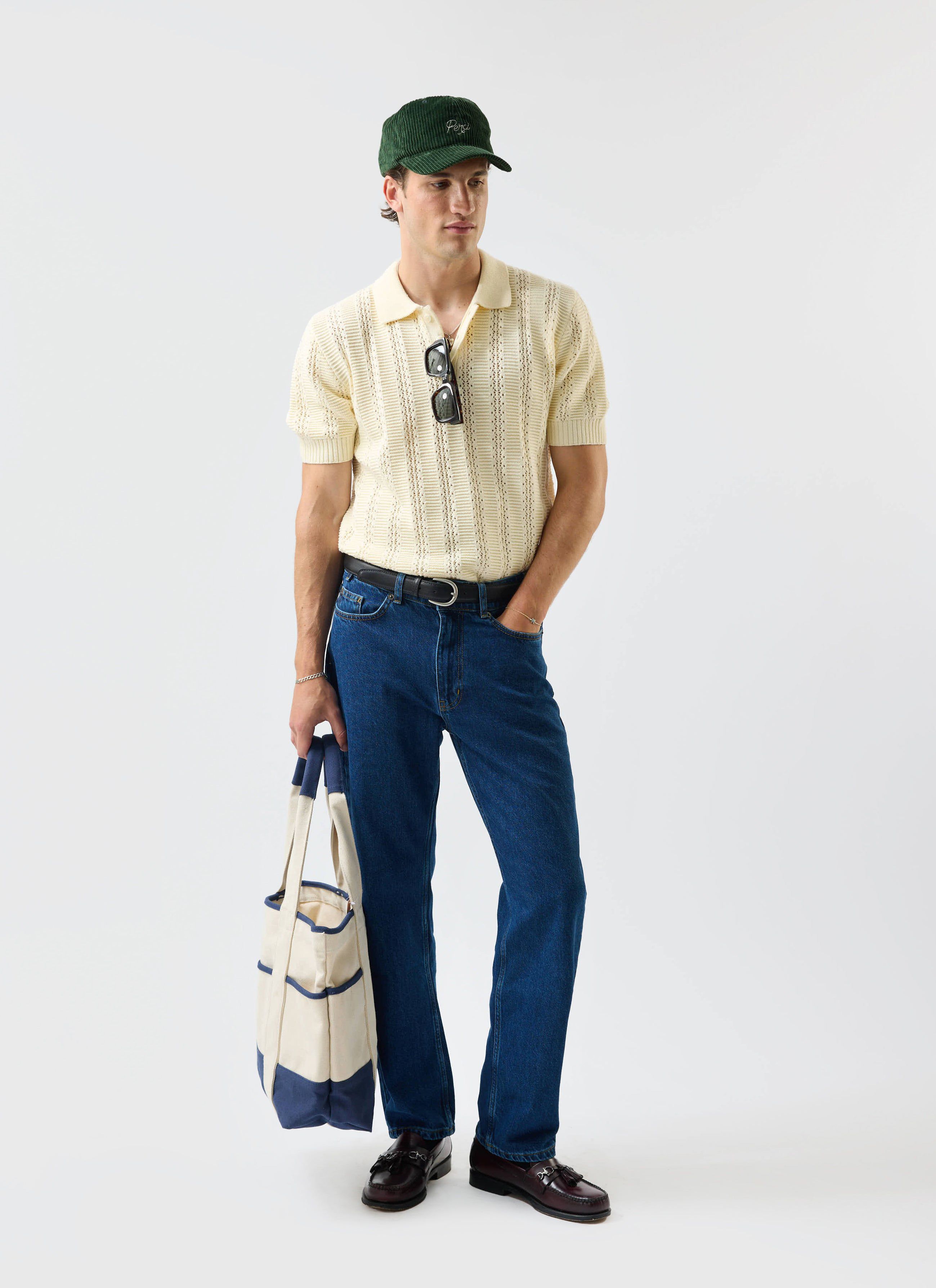 A model styled in the Percival Addendum Knitted Polo Shirt in ecru cream, paired with blue jeans and a green corduroy cap.