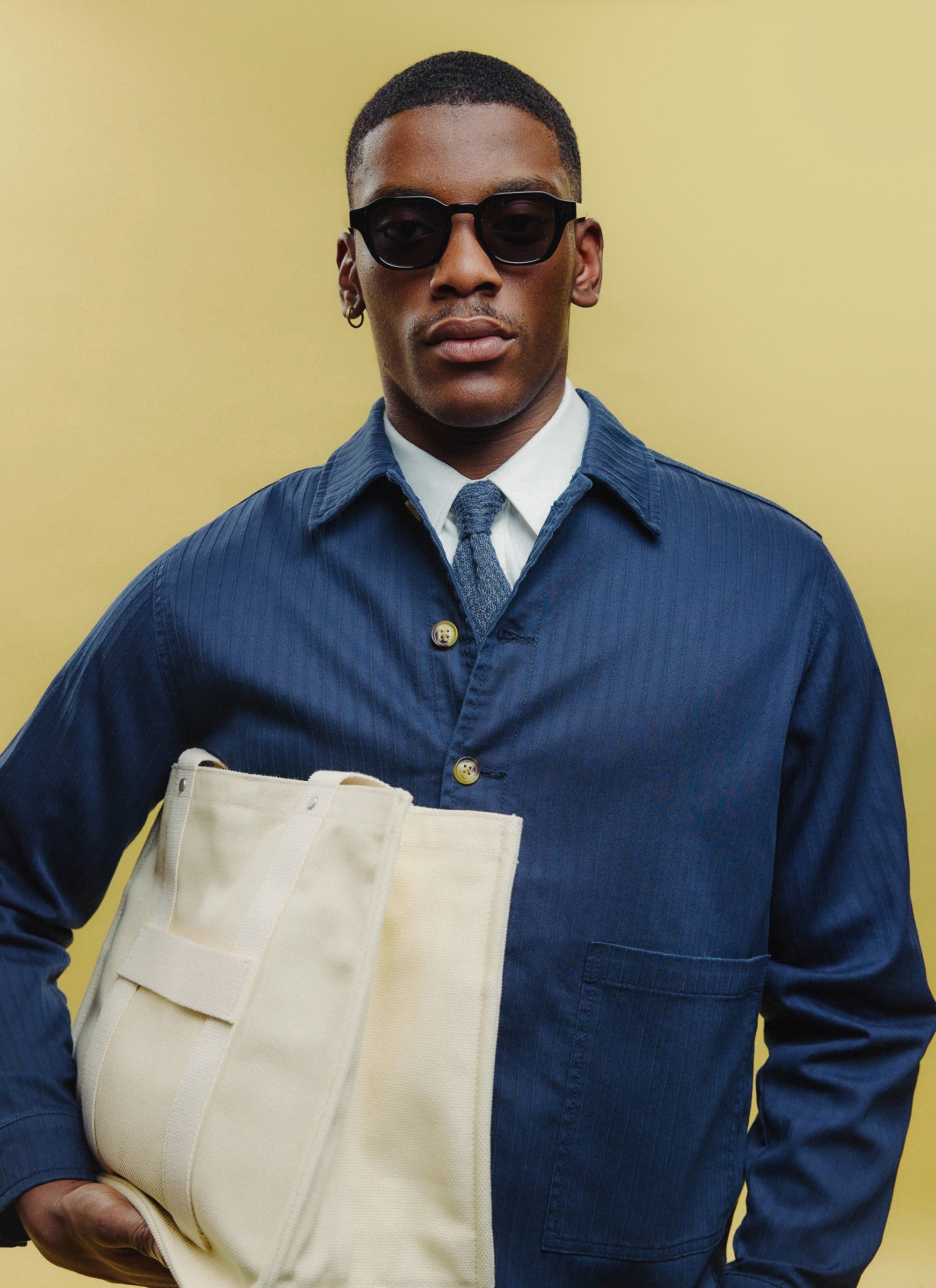 A model wears the black Percival sunglasses with a navy jacket, white shirt, blue tie, and holds a cream tote bag.