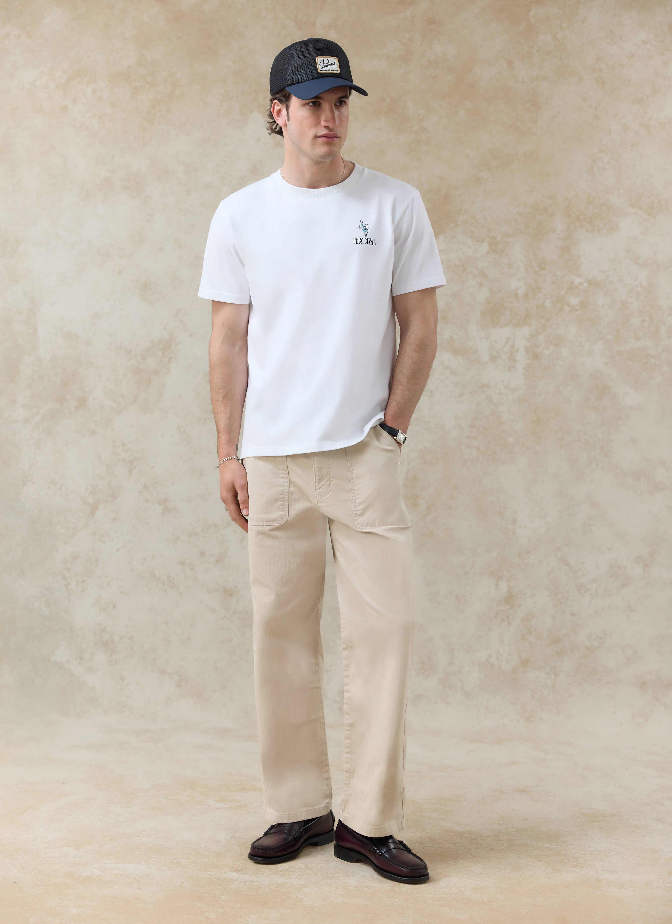 White organic cotton Percival Walker T Shirt with a chest graphic, styled with beige trousers and a navy cap.