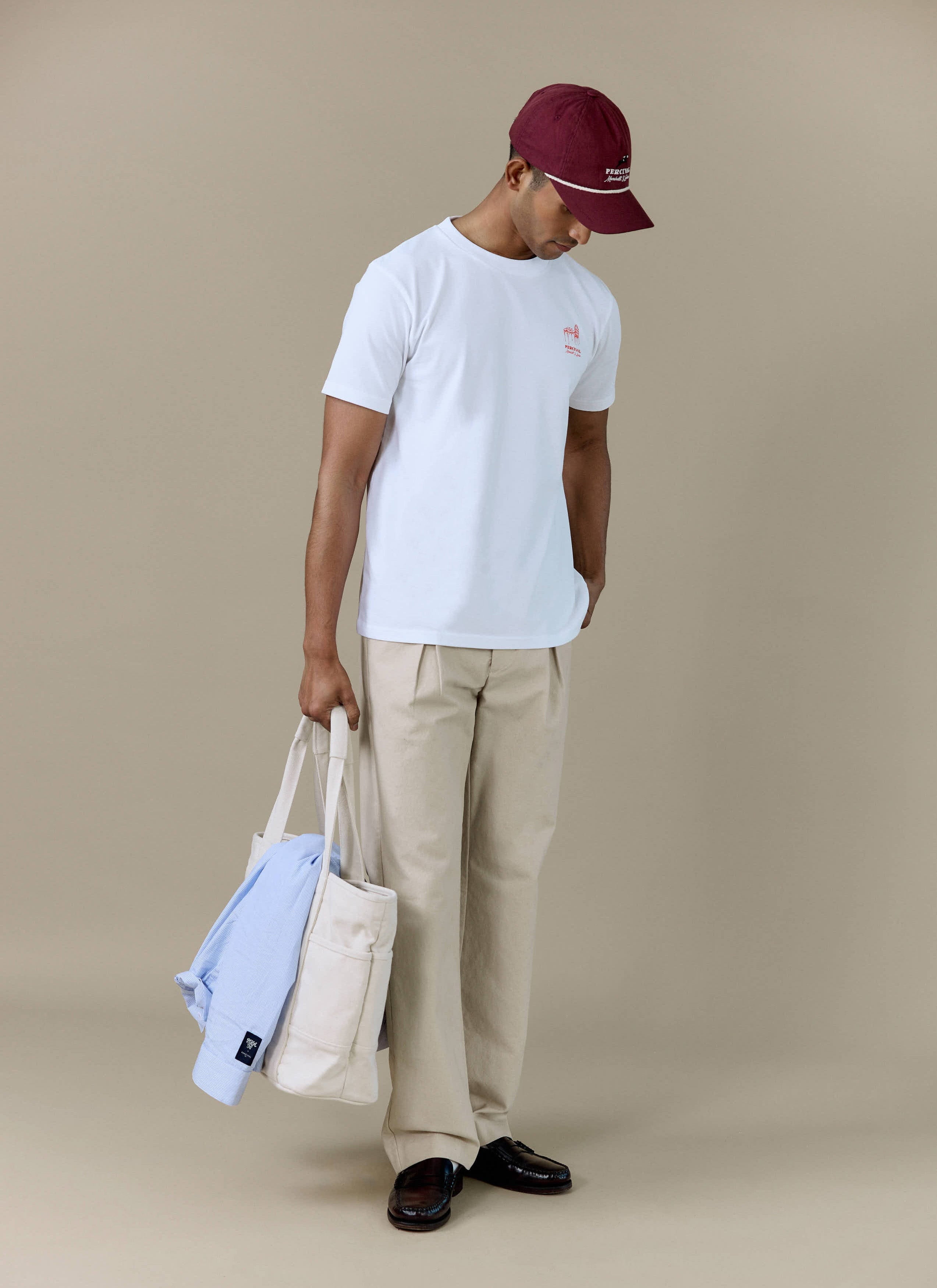 A model wearing the white Perci Fine Wines T Shirt by Percival, styled with beige trousers and a burgundy cap.
