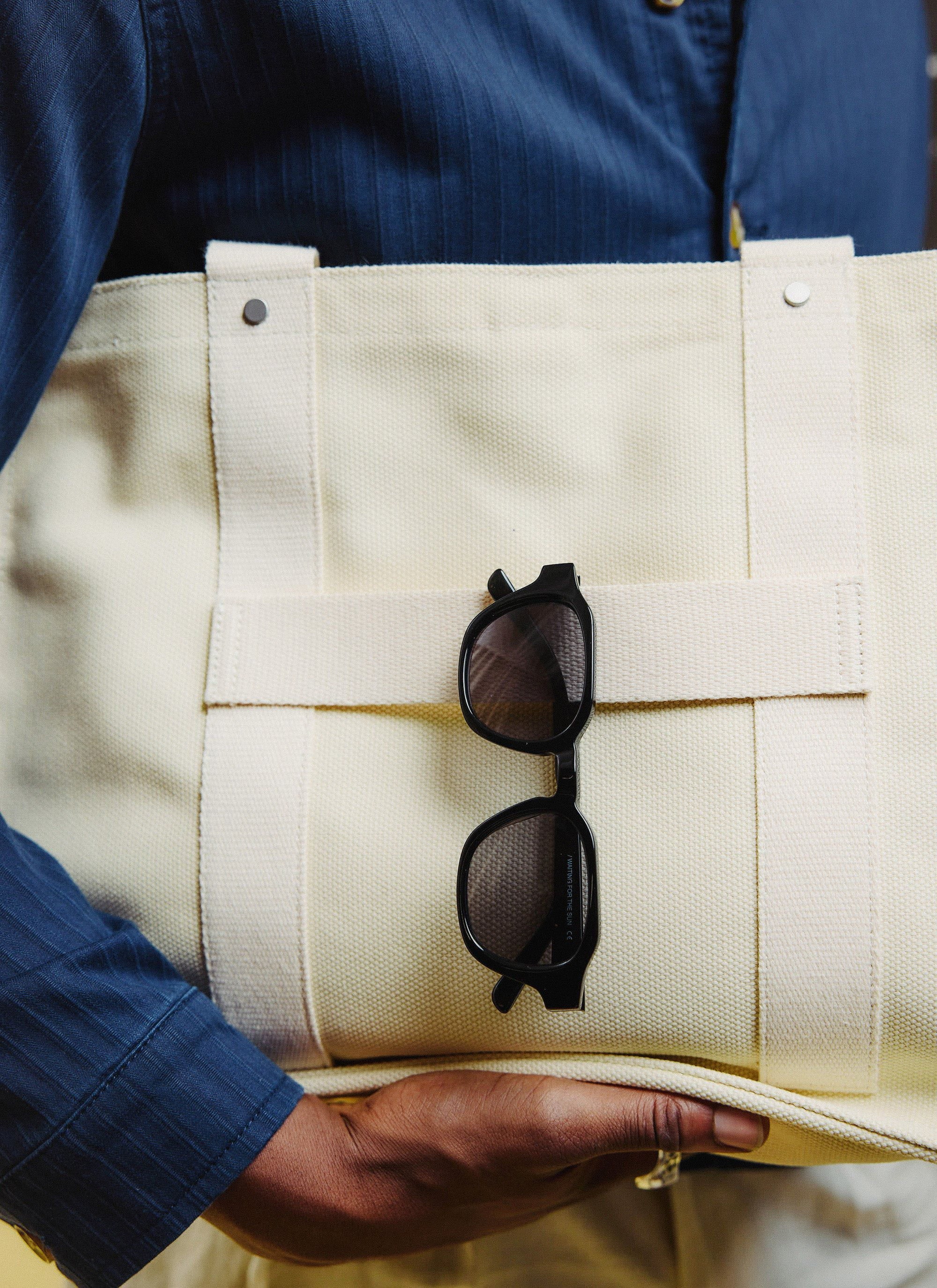The Percival black sunglasses, with polarised lenses, tucked into the pocket of a cream canvas tote bag.