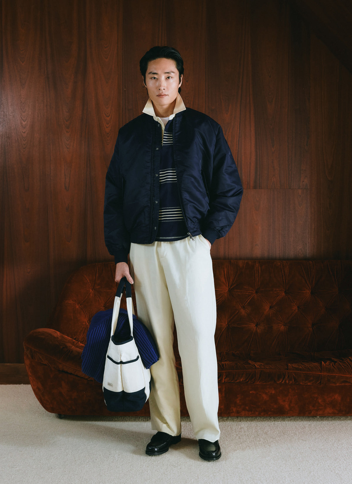 A model wears the Percival midnight blue nylon bomber jacket with a patterned polo and tote bag against a wood panel wall.