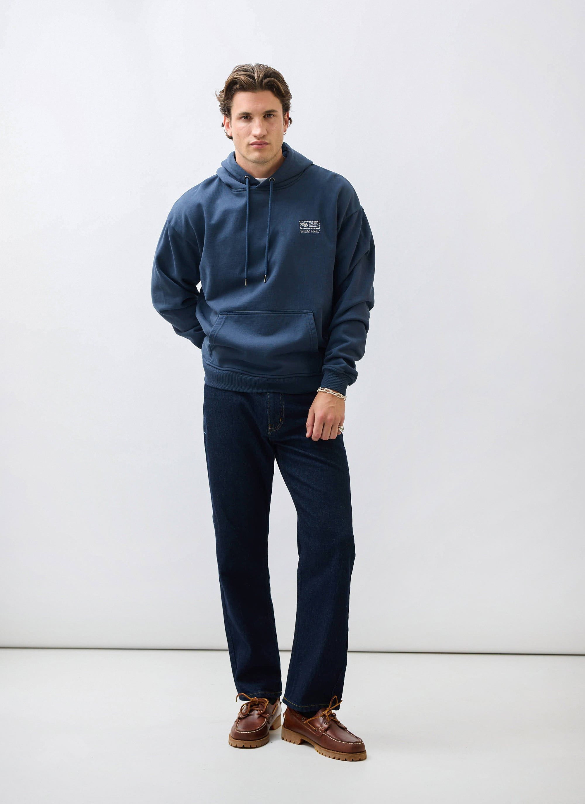 A model wears the Percival 'This Book Belongs To' hoodie in navy, styled with dark denim jeans and brown boat shoes.
