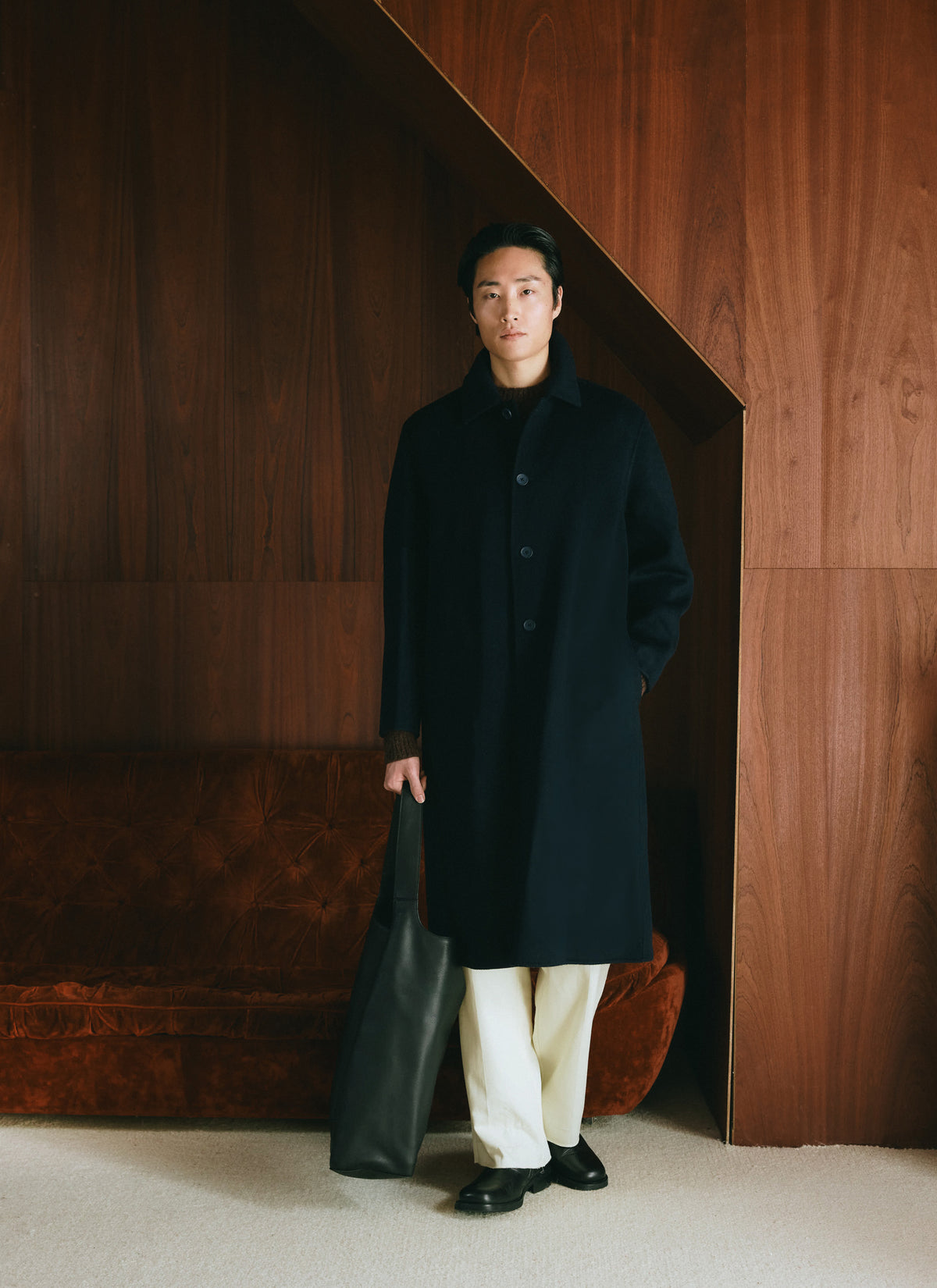 A model wearing the Percival navy wool Minnesota overcoat and holding a bag in a wood-panelled room.