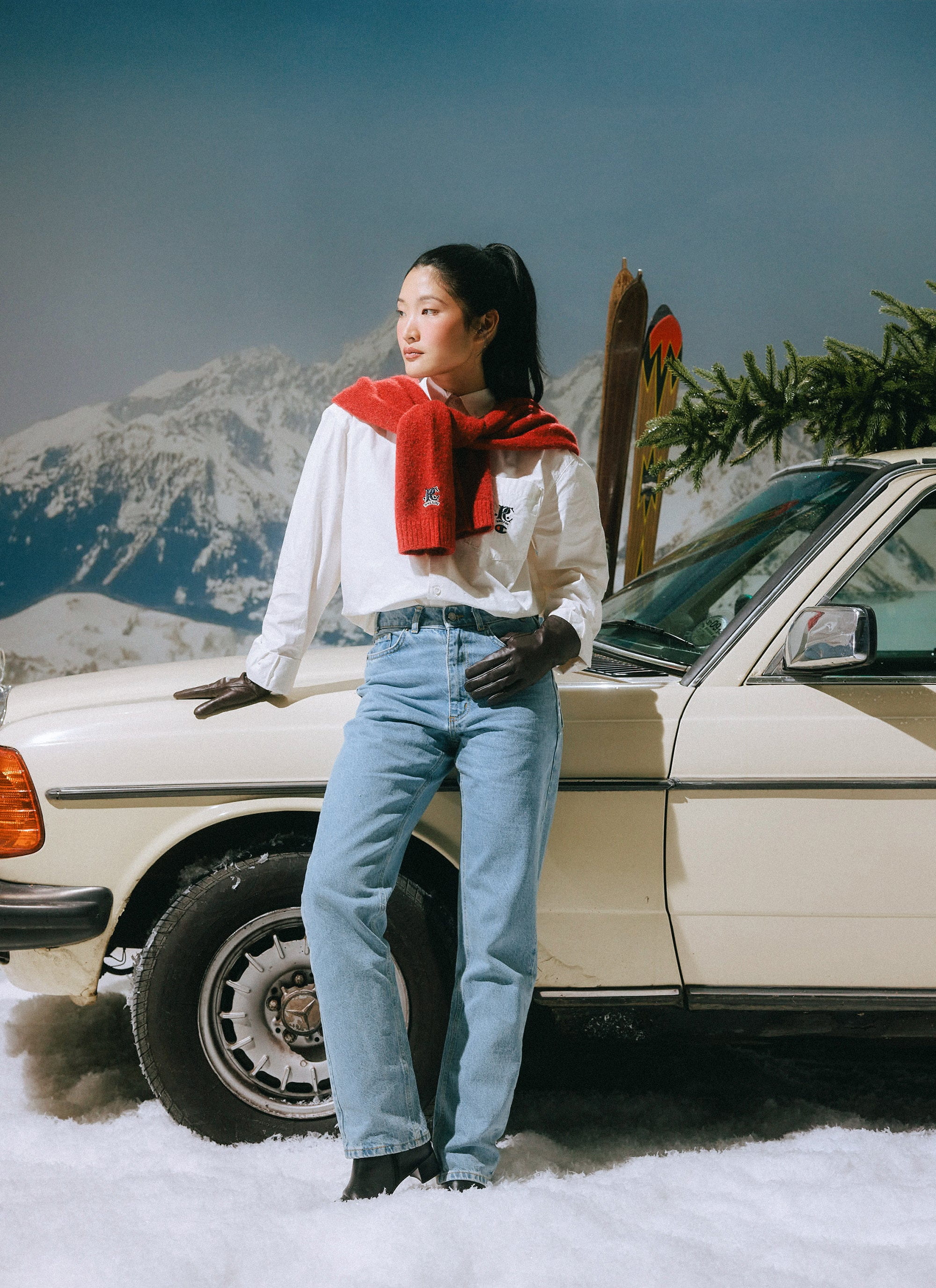 A model wears the ecru Champion x Percival Oxford shirt, leaning against a vintage car in a snowy, après-ski setting.