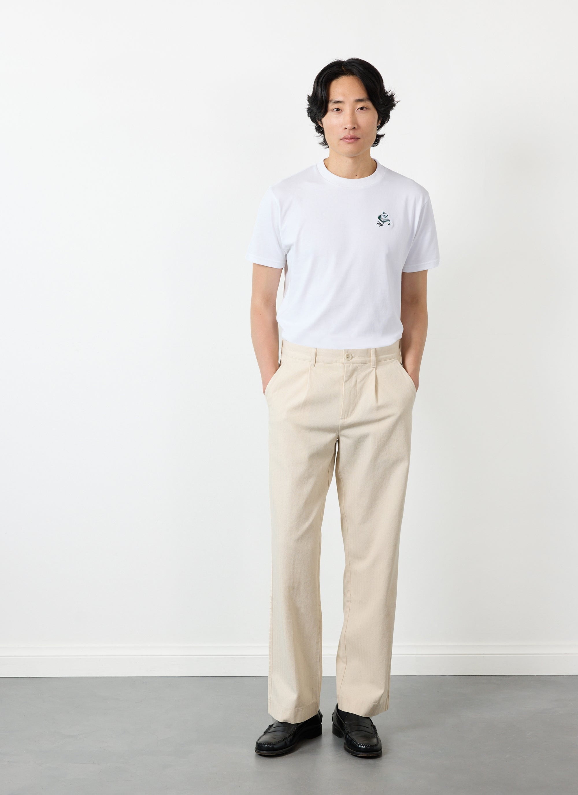 A model wears Percival's Program Pleated Trousers in ecru with a white t-shirt and black loafers.