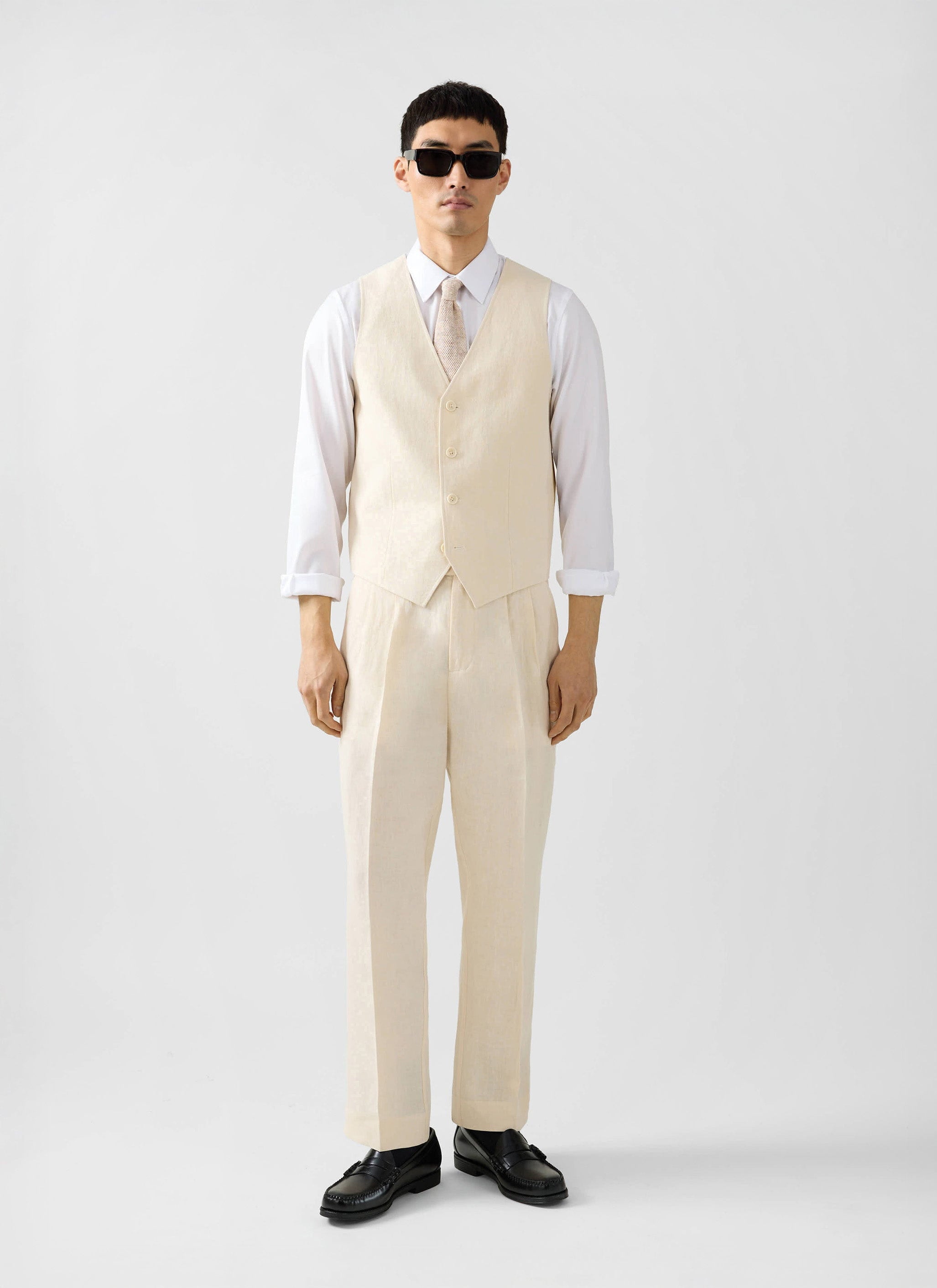 A model wears the Percival cream linen waistcoat and matching trousers, styled with a white shirt, tie and sunglasses.
