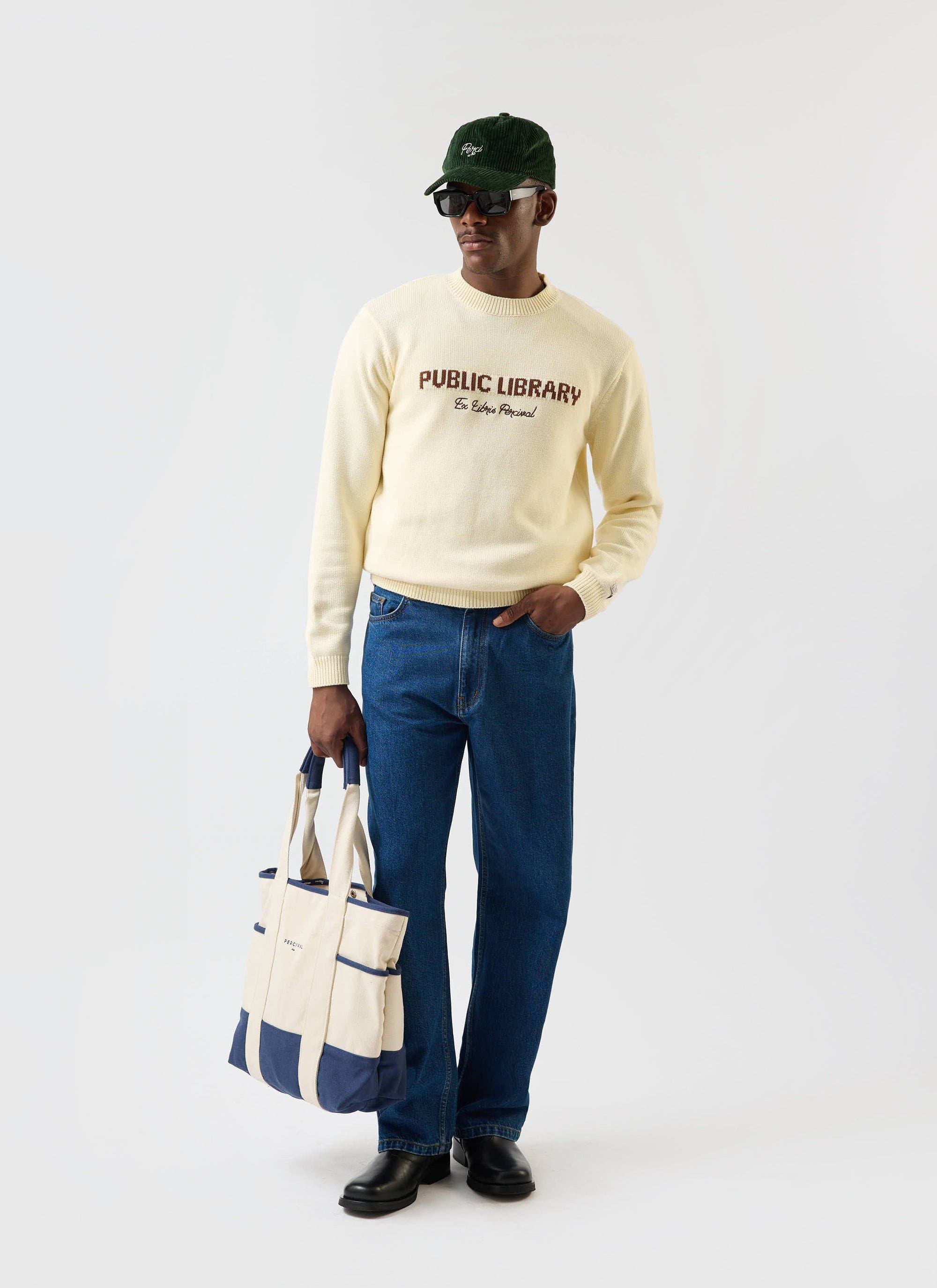 A model wears the ecru cream Public Library knitted jumper from Percival, styled with blue jeans, a tote bag and a green cap.