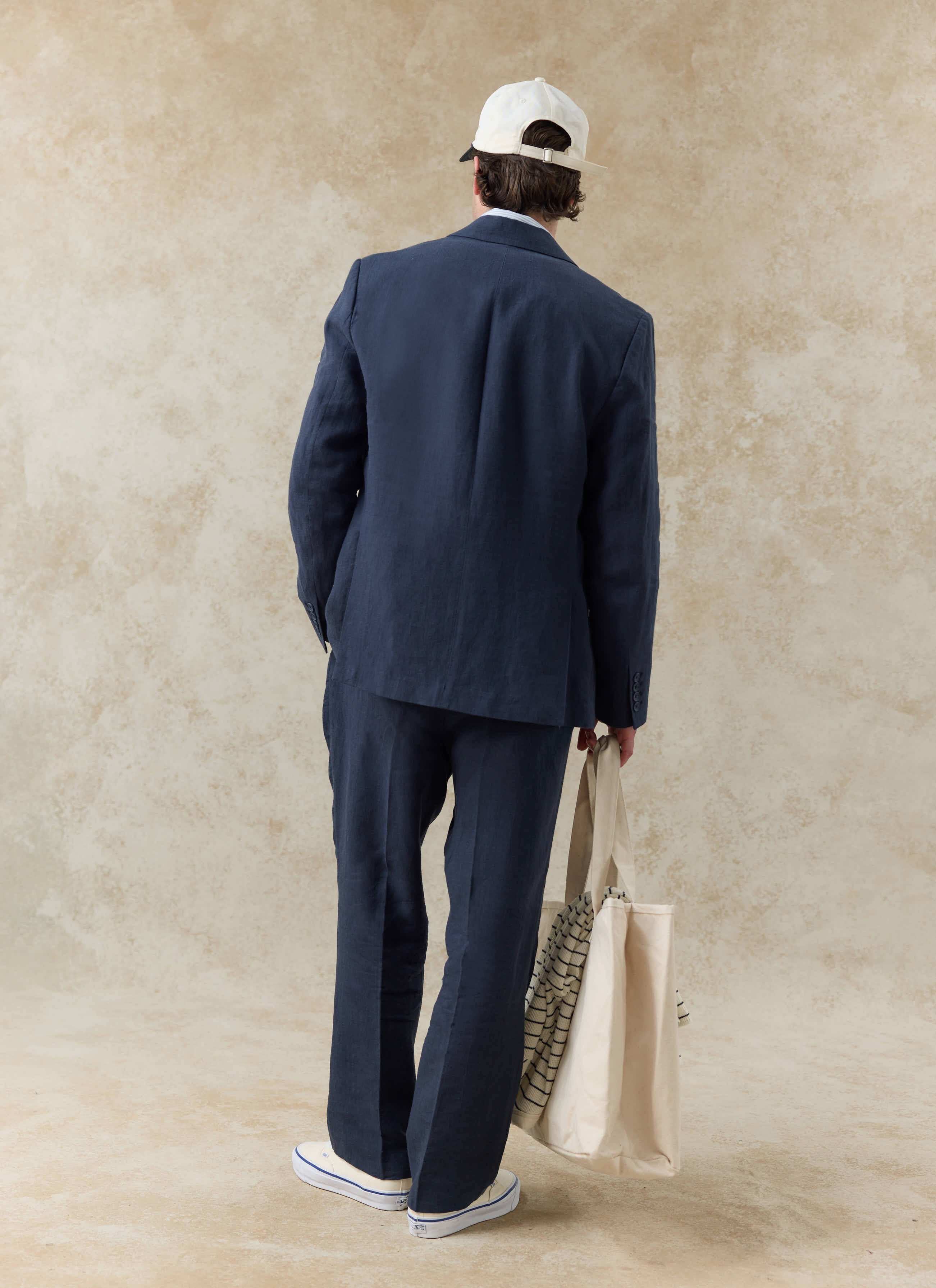 Rear view of a model wearing the navy linen Percival suit, featuring relaxed fit pleated tailored trousers.