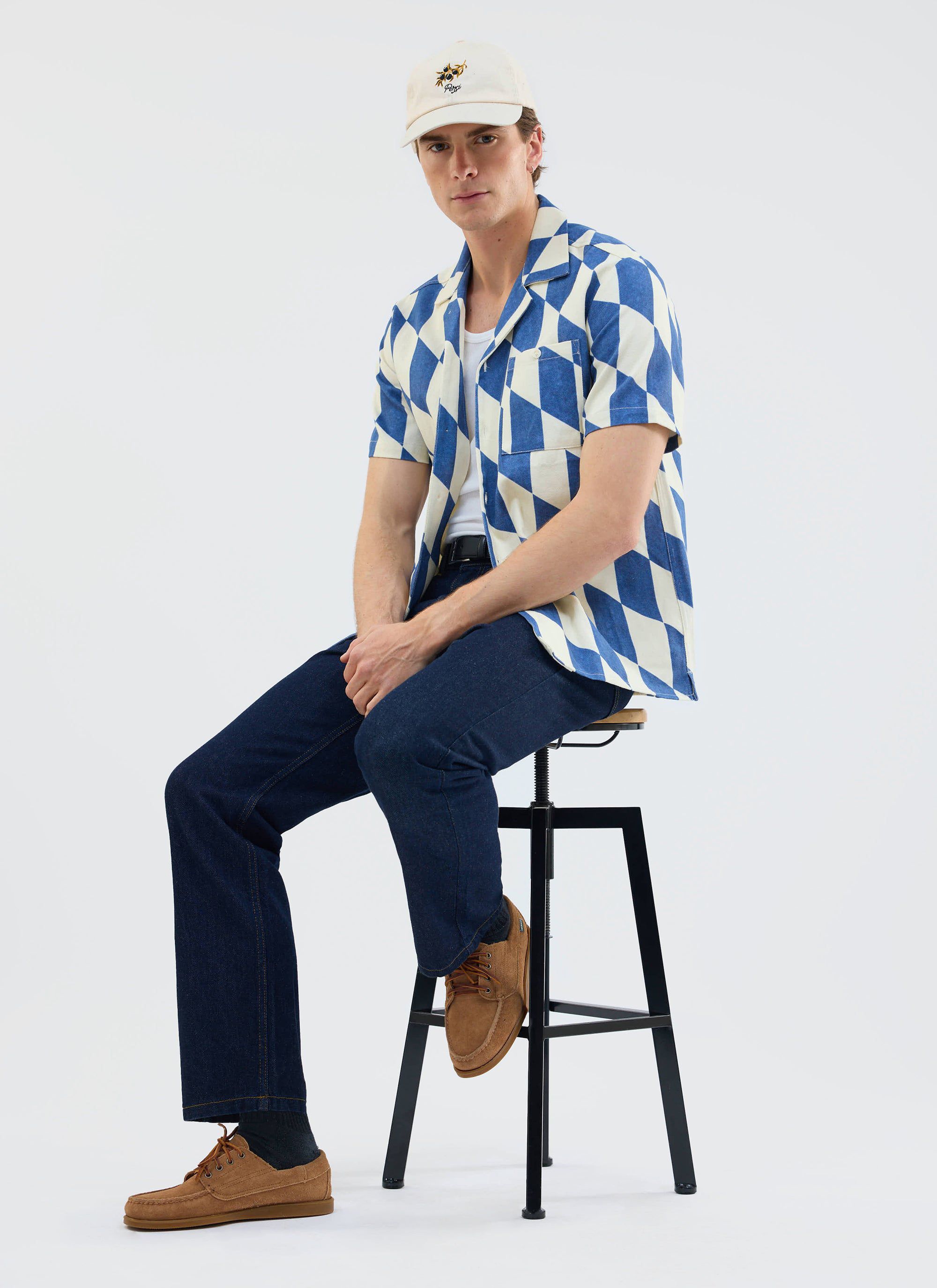 A model wears the Percival Market Diamond Cuban shirt in ecru and navy with dark jeans and a cap while sitting on a stool.