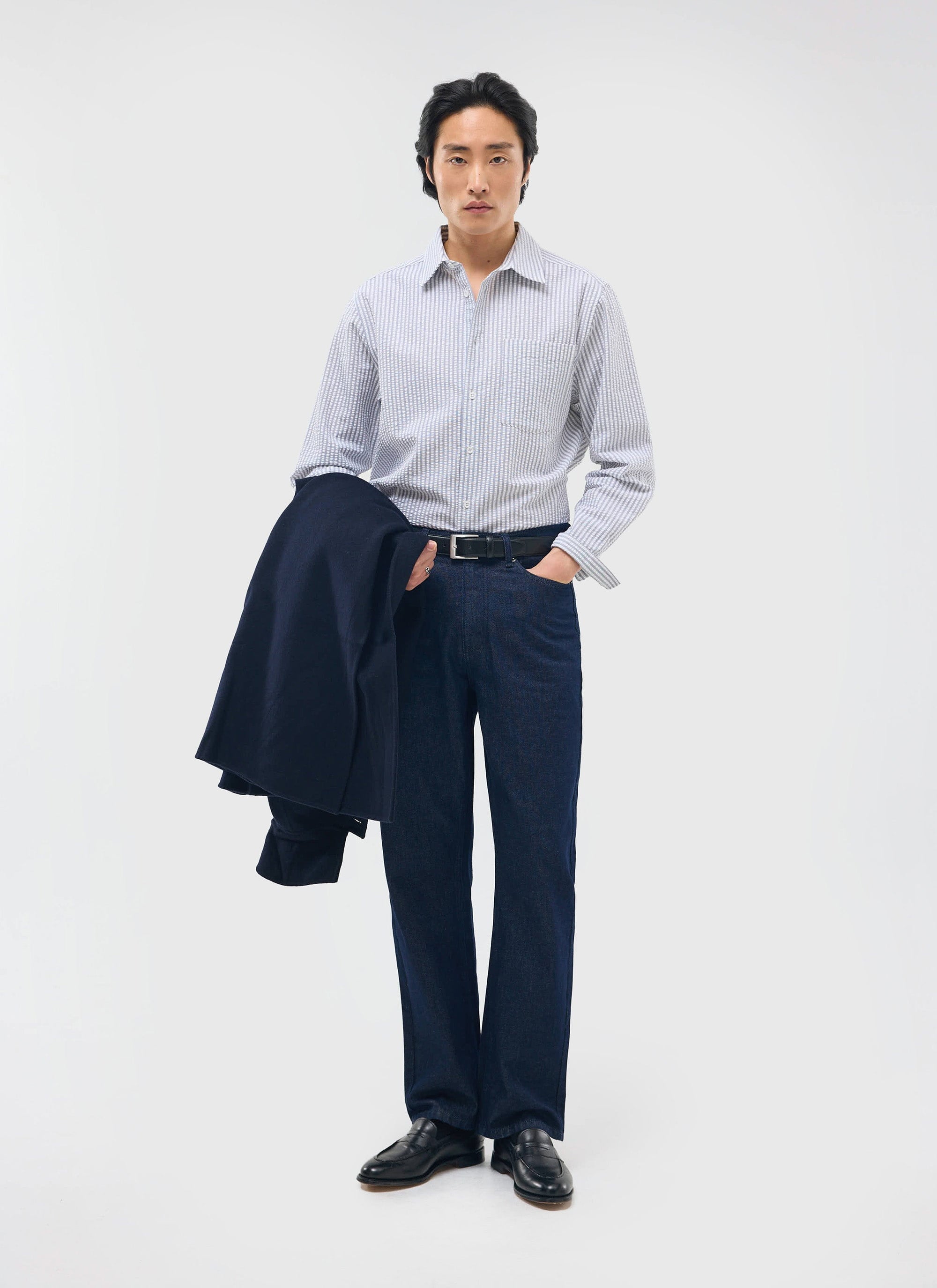 A model in a studio setting wears the Percival Seersucker Stripe Pearce Shirt in grey, with dark jeans and black loafers.