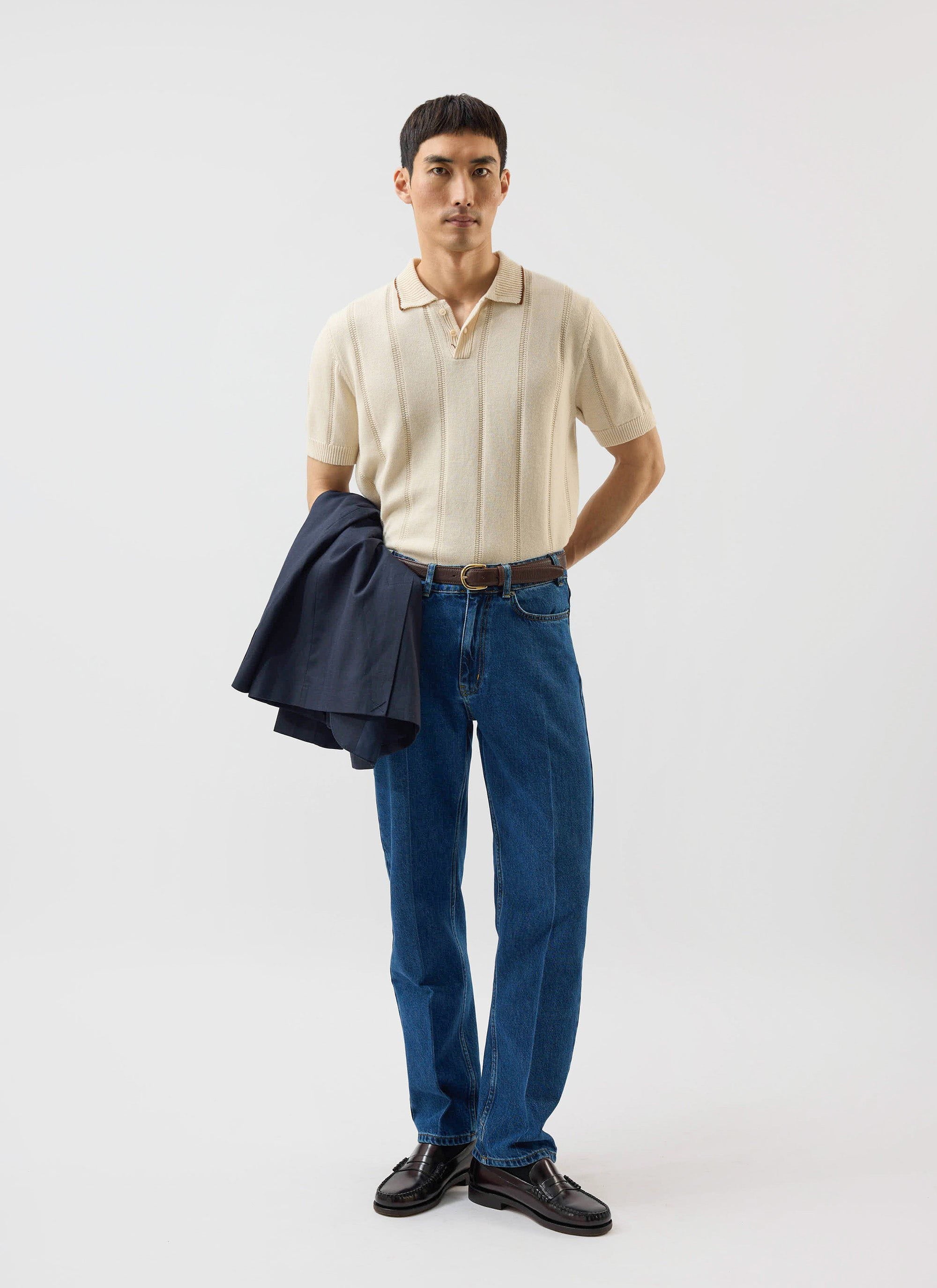 A model wears the ecru Percival x Warren Knitted Polo, styled with blue jeans and holding a navy blazer.