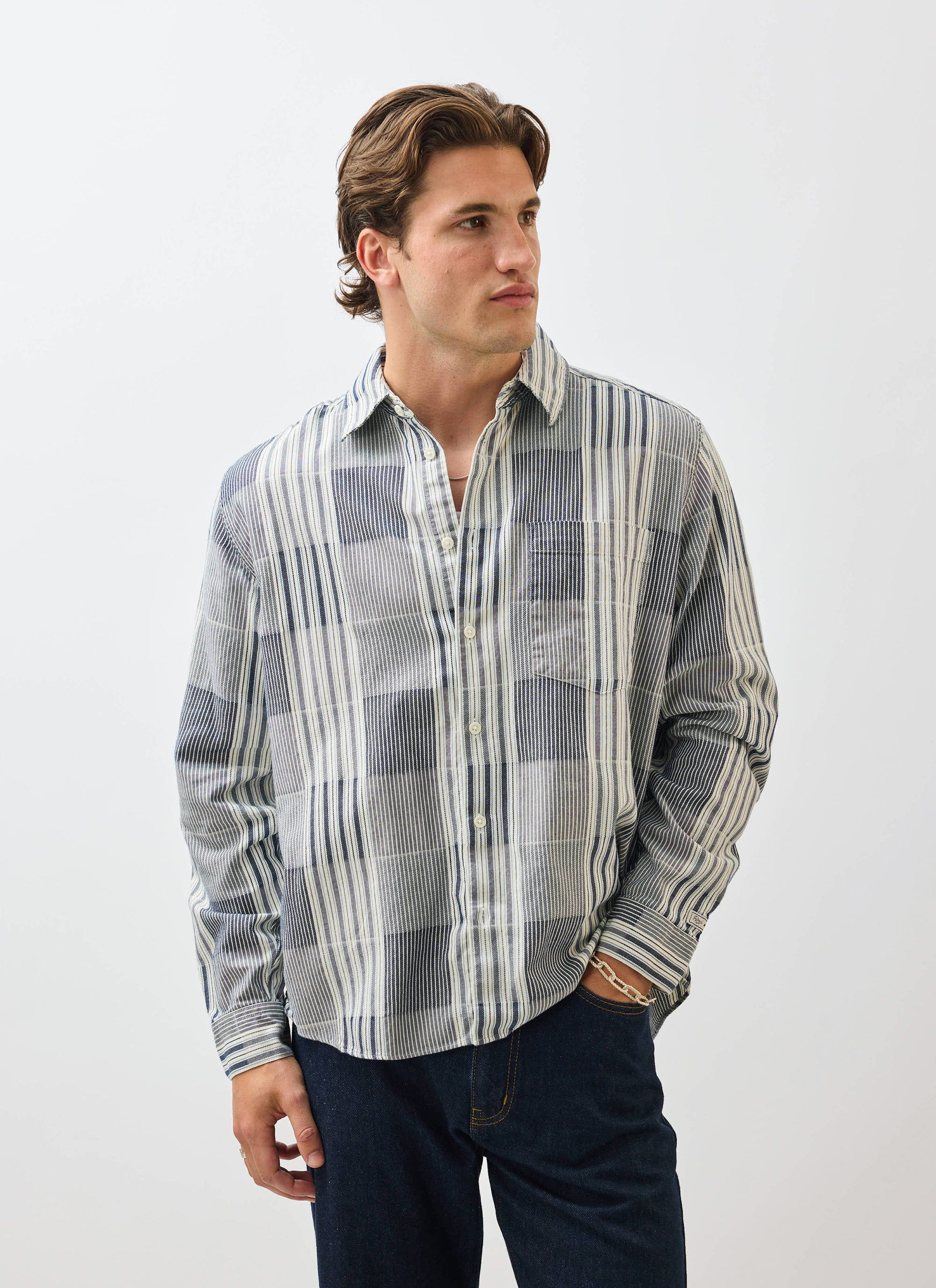 A man models the Percival Epigraph Patchwork Pearce Shirt in blue, worn with dark jeans against a white background.
