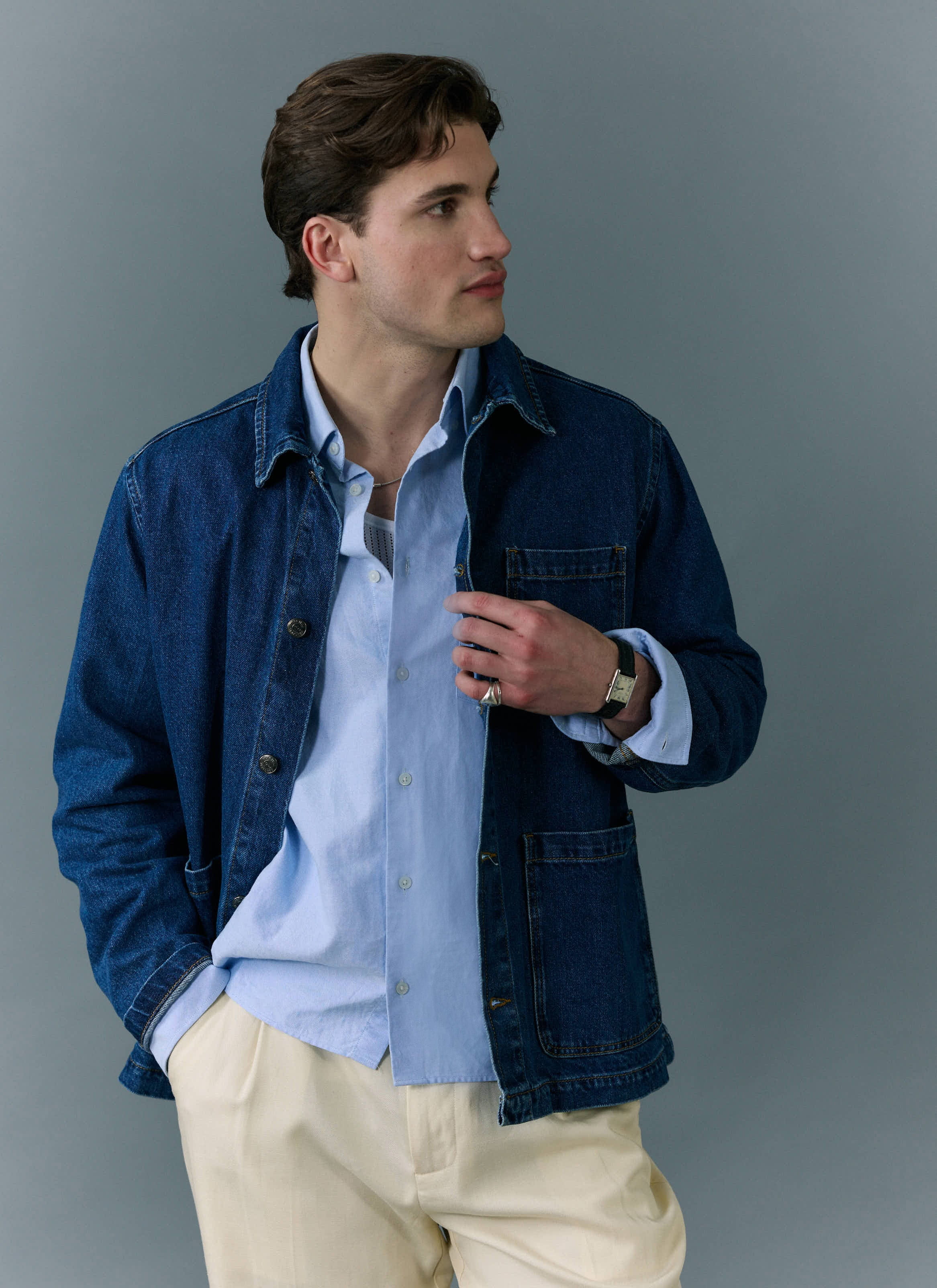 A man models the Percival Gove Chore Jacket in mid-wash blue, styled over a light blue shirt and cream trousers.