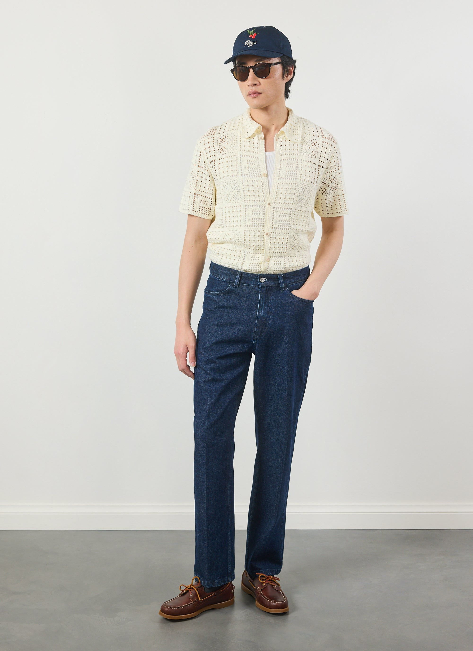 A model wears the Percival Crochet Groundsman Shirt in ecru, styled with dark denim jeans, a navy cap and sunglasses.