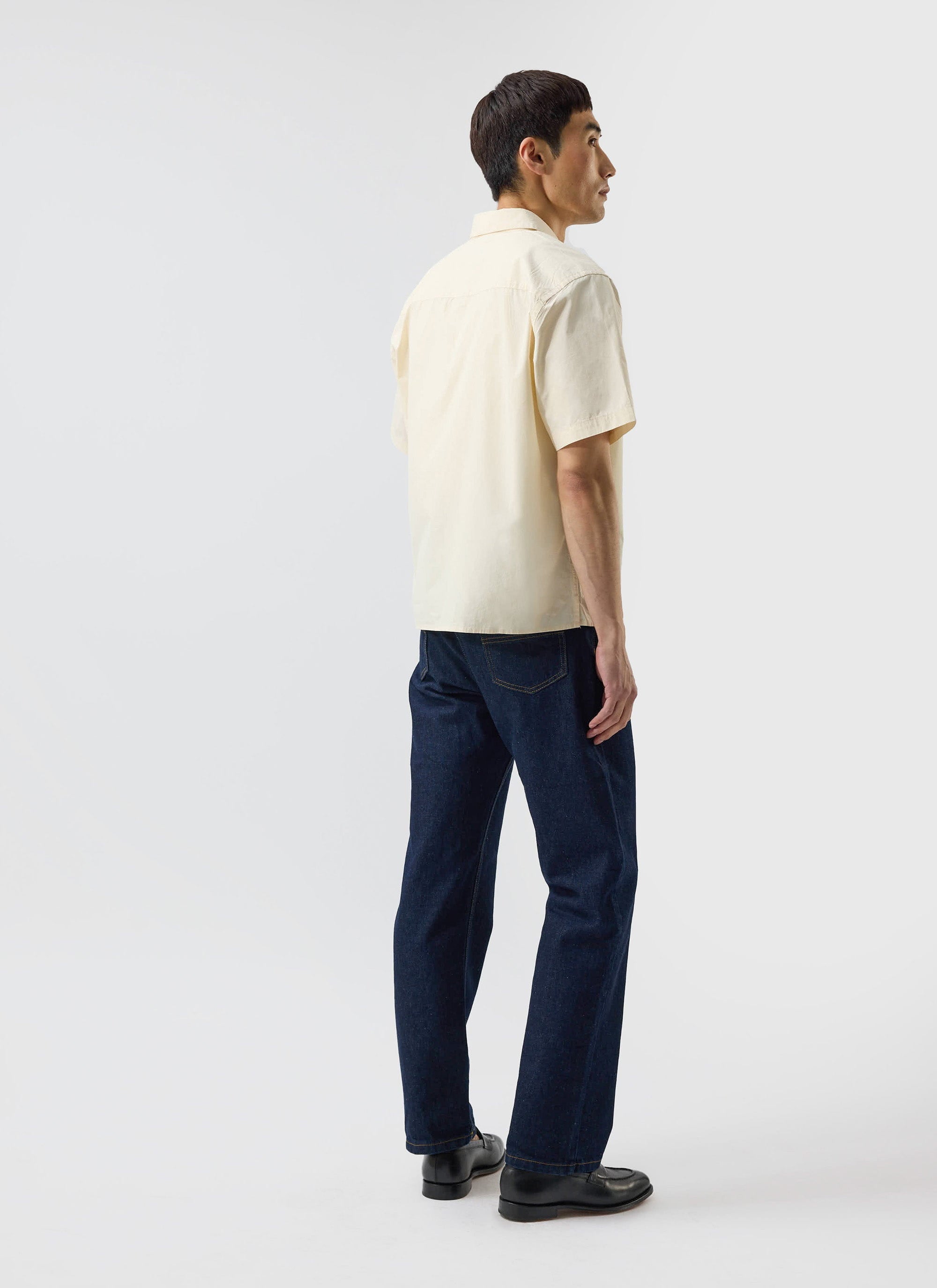 A man models the Percival Boxy Cropped Clerk Shirt in ecru with dark jeans, seen from a three-quarter back view.