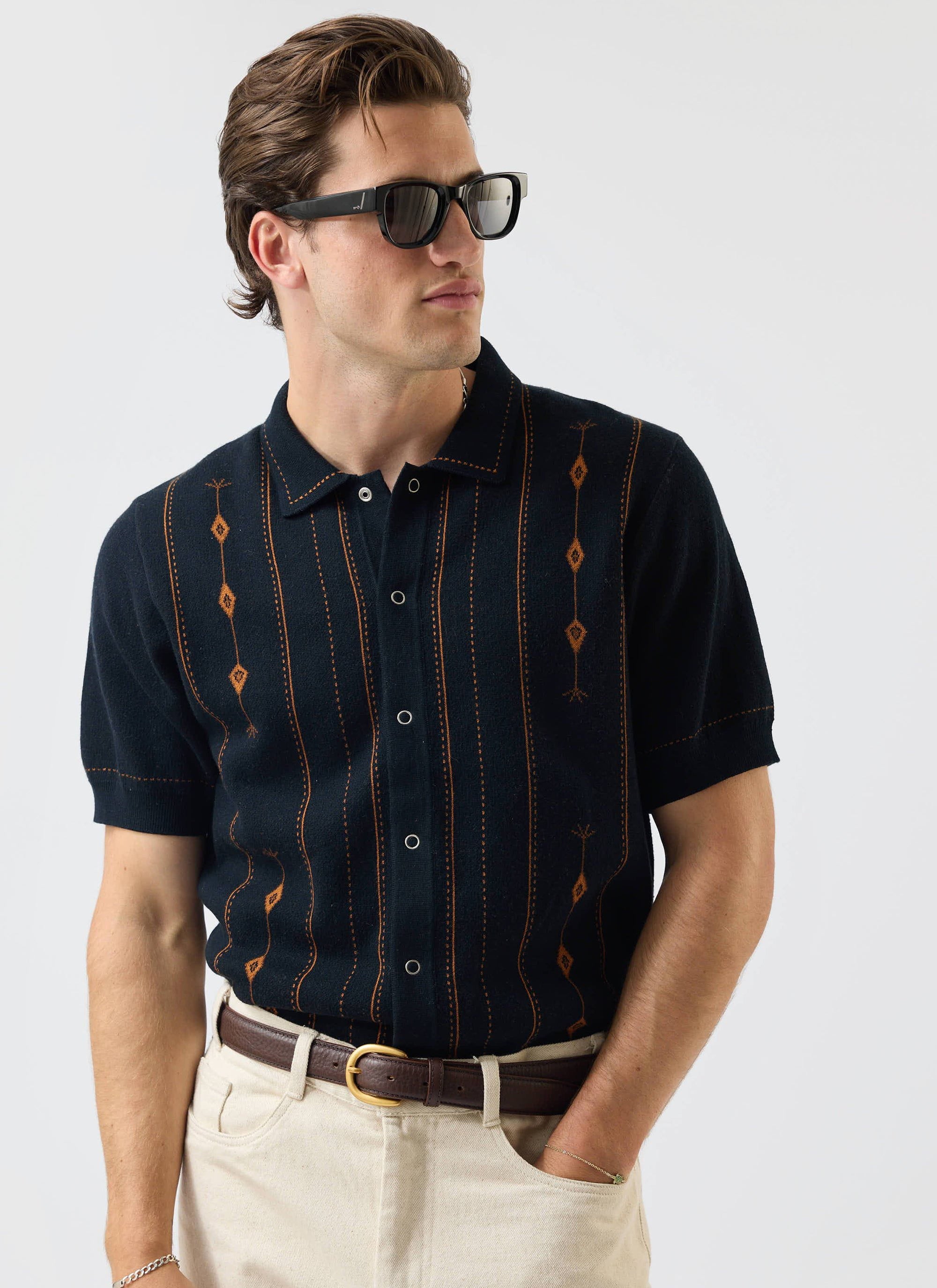 A man models the Percival Folio Knitted Shirt in navy, featuring a tobacco jacquard pattern, styled with cream trousers.