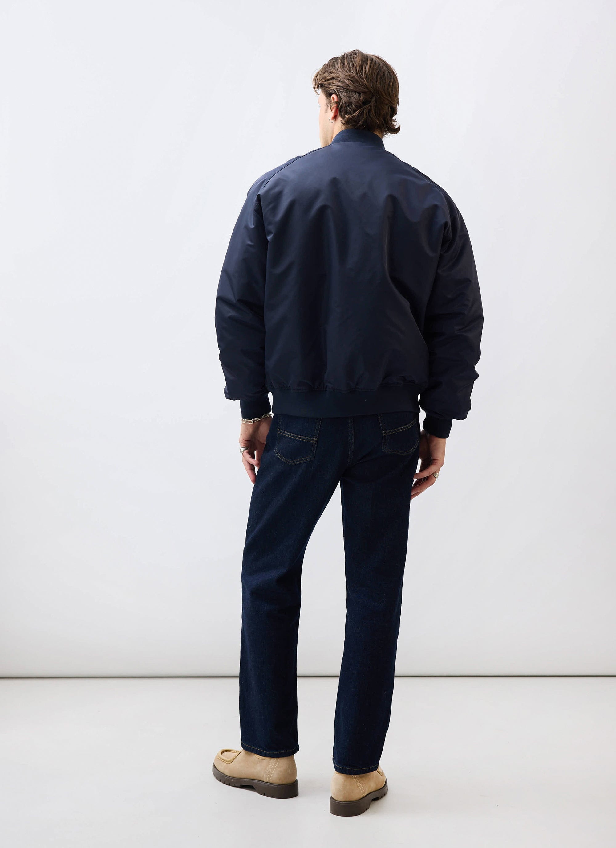 A model, seen from behind, wears the Percival nylon bomber jacket in midnight navy with dark denim jeans.