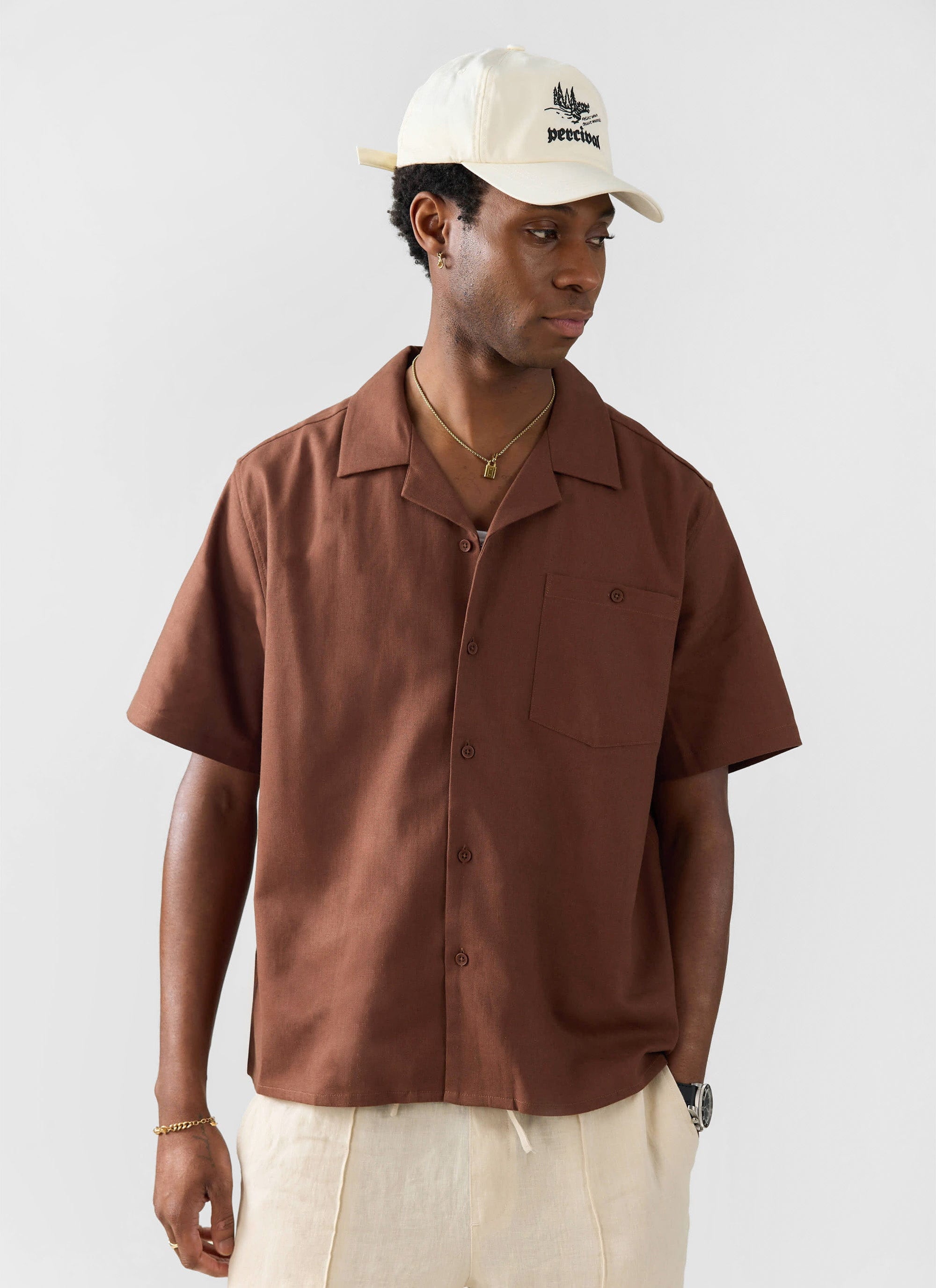 The Percival Short Sleeve Cuban Shirt in brown linen, styled on a model with a white cap and cream trousers.