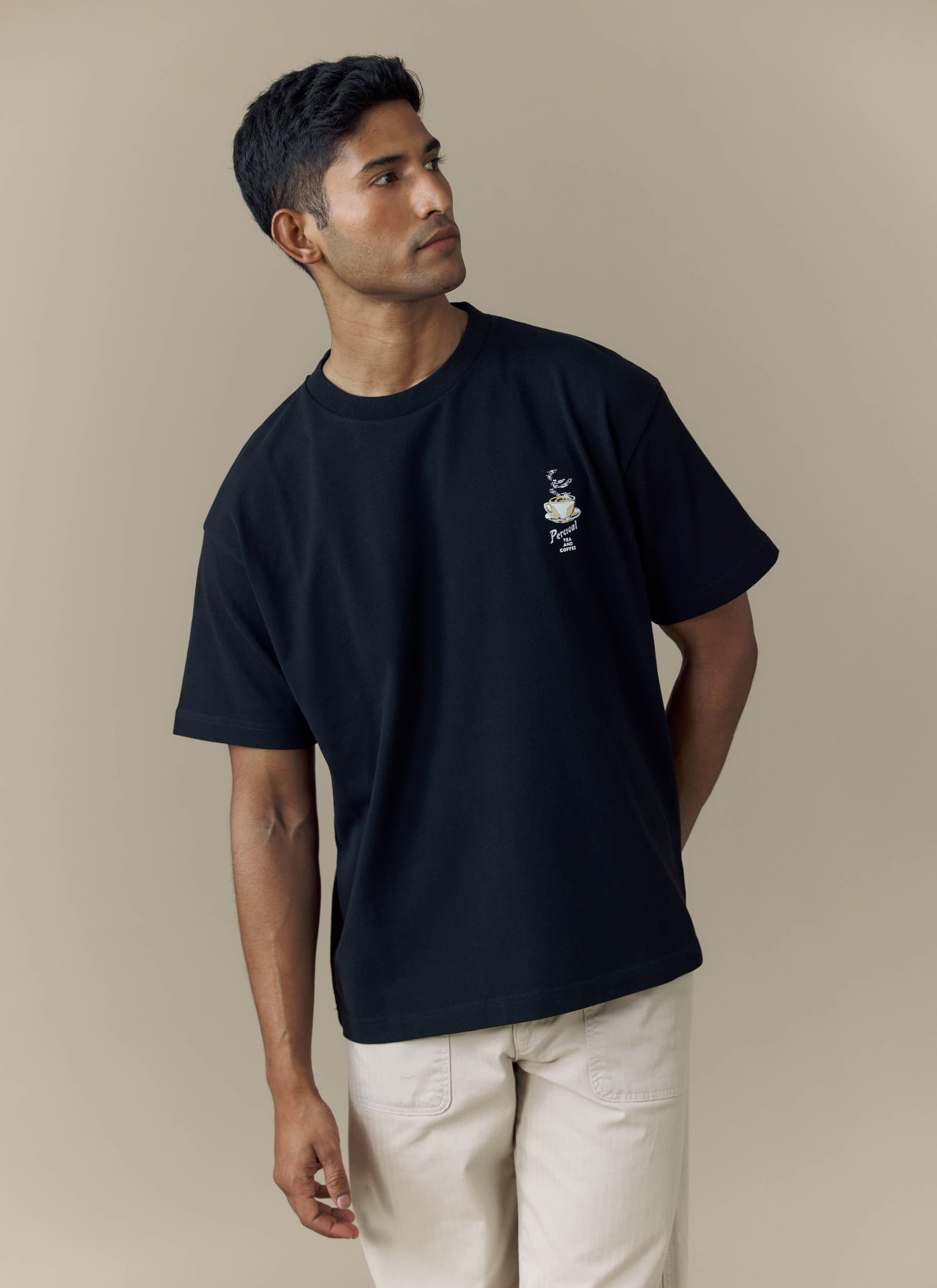 A model wears the black Percival Grill Oversized T-Shirt with its chest embroidery, styled with light-coloured trousers.