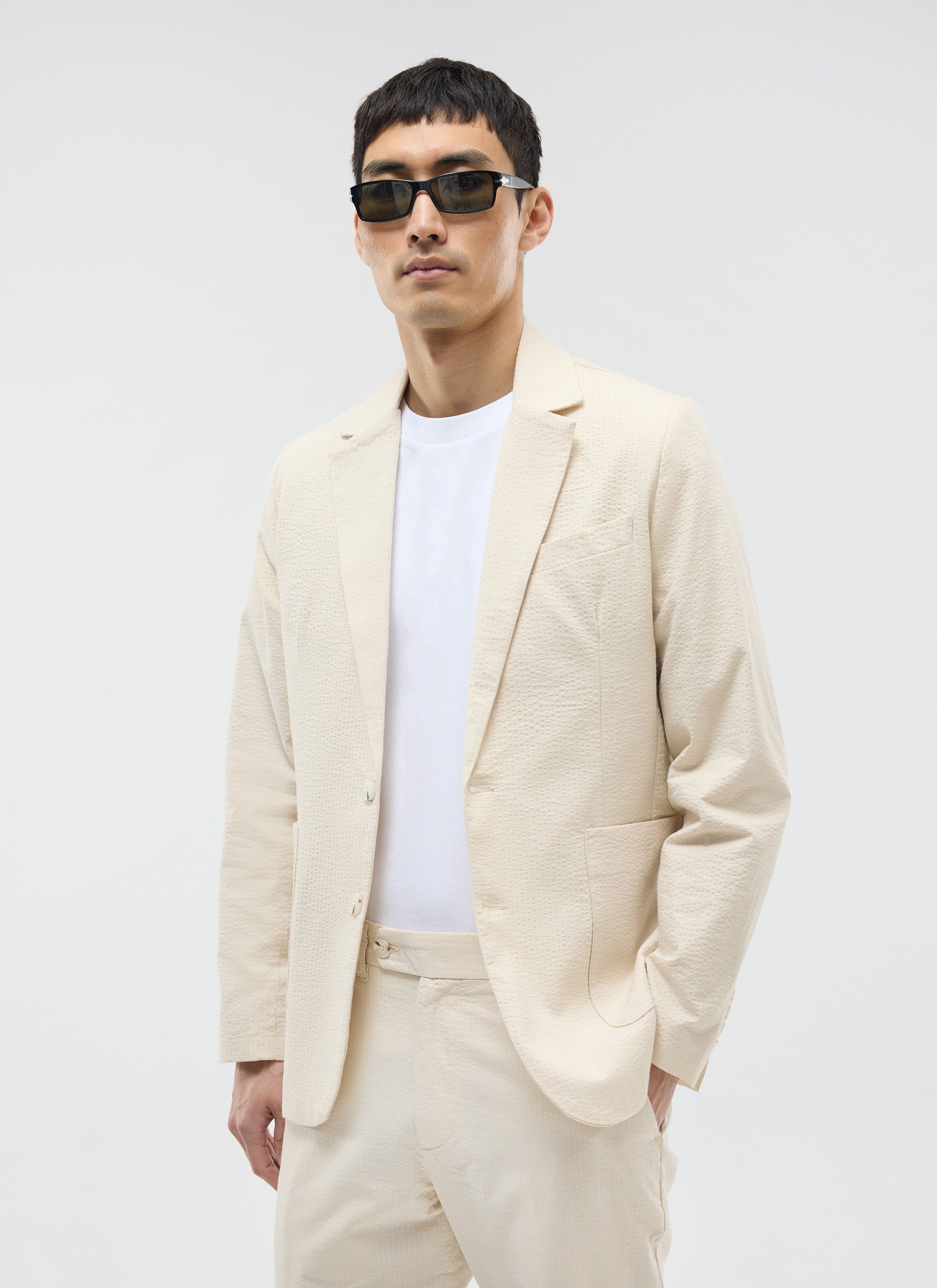 A model wears the Percival Tailored Seersucker Blazer in cream, styled with a white t-shirt and black sunglasses.
