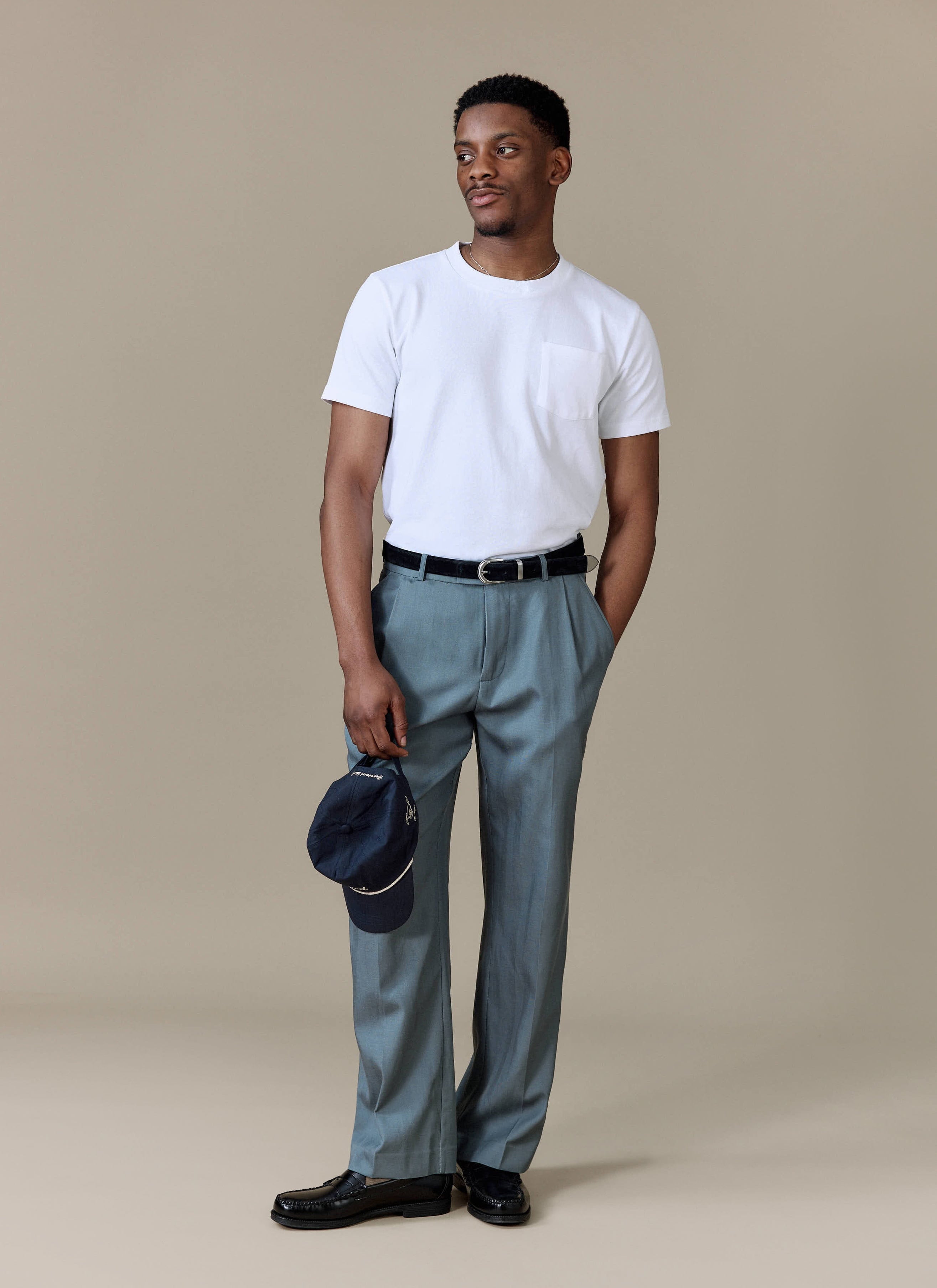 A model wears the Percival Pleated Leg Trousers in Slate Blue, styled with a white t-shirt and black loafers.