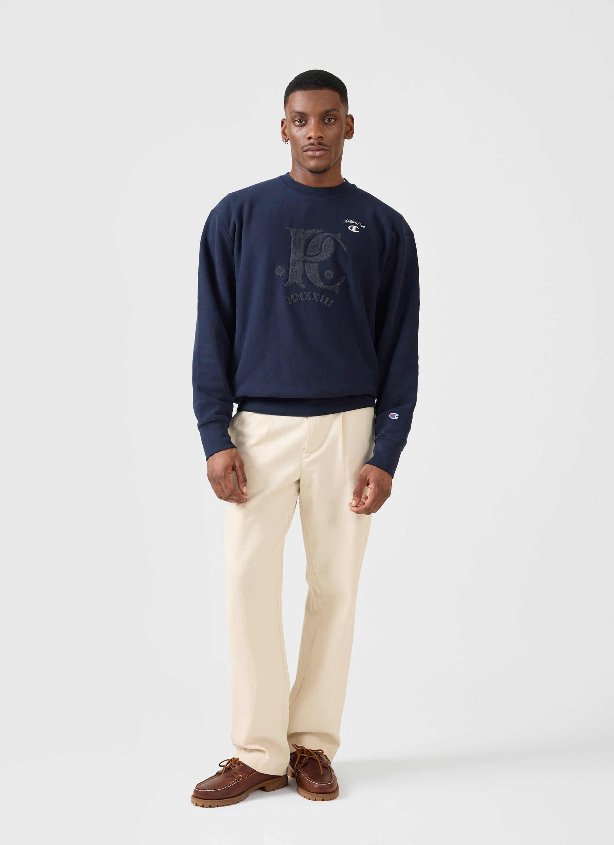 A model wears the navy Champion x Percival Chenille Patch Jersey Sweatshirt, paired with cream trousers.