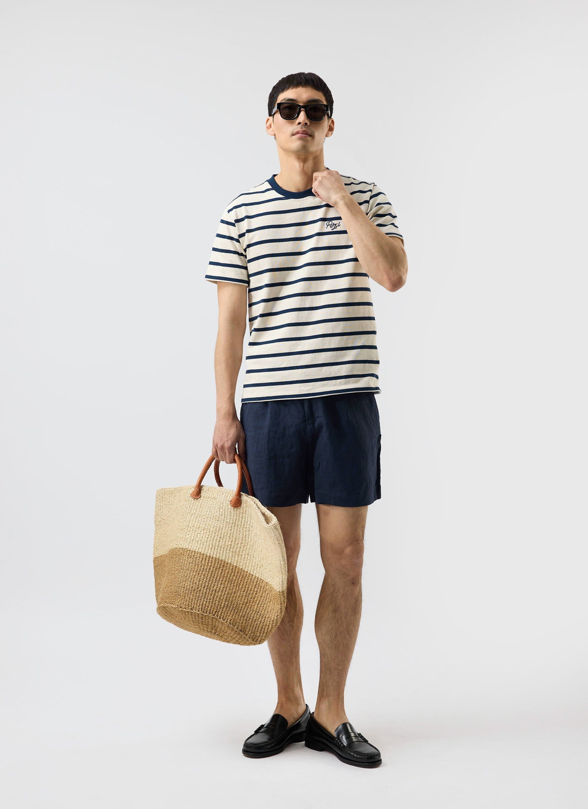 A model wears the Percival Breton Stripe T Shirt in ecru and navy, styled with navy shorts, sunglasses, and a woven tote bag.