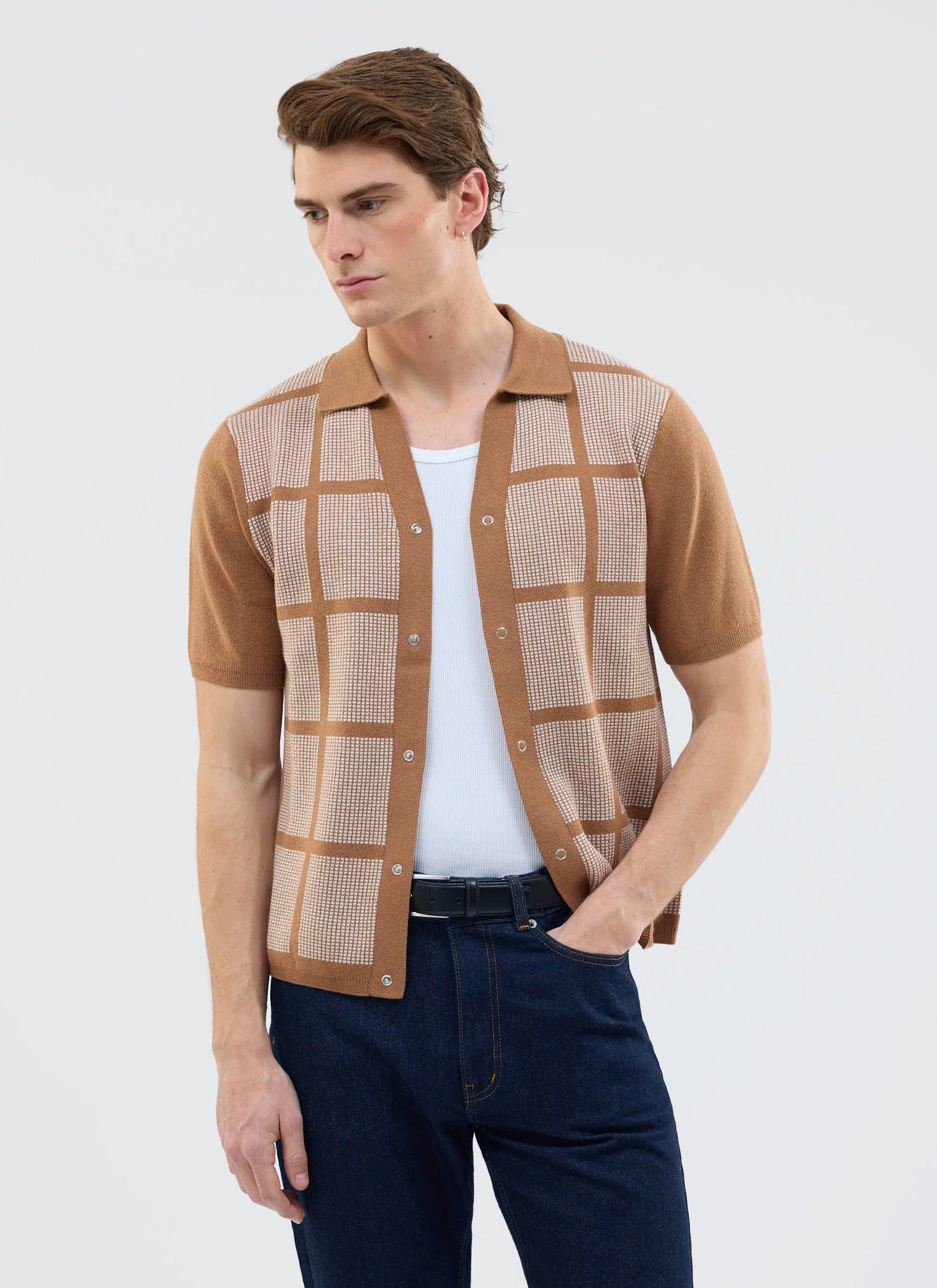 A man models the tobacco brown Percival Costa Knitted Shirt, with a square pattern, worn open with a white vest and jeans.