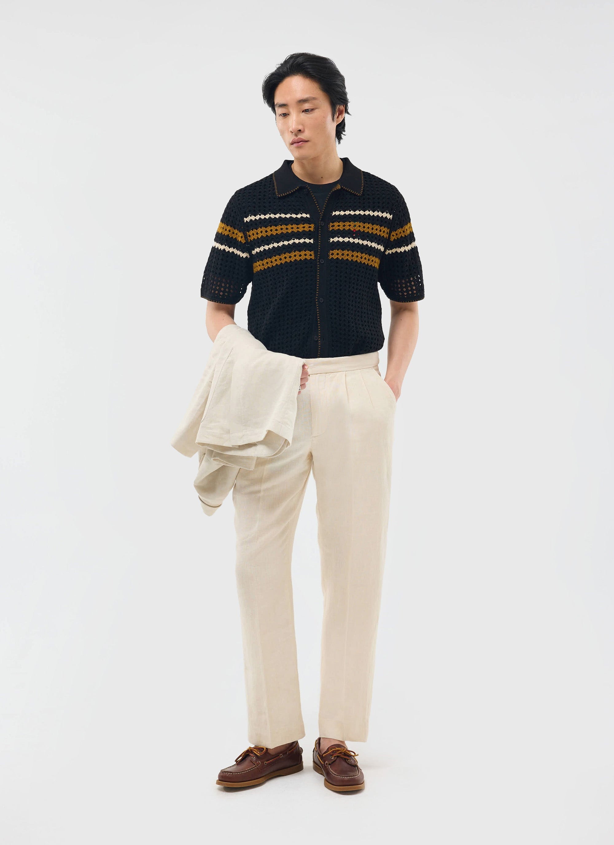 A model wears the black Percival Pesaro Knitted Shirt, featuring mustard and white stripes, with cream trousers.