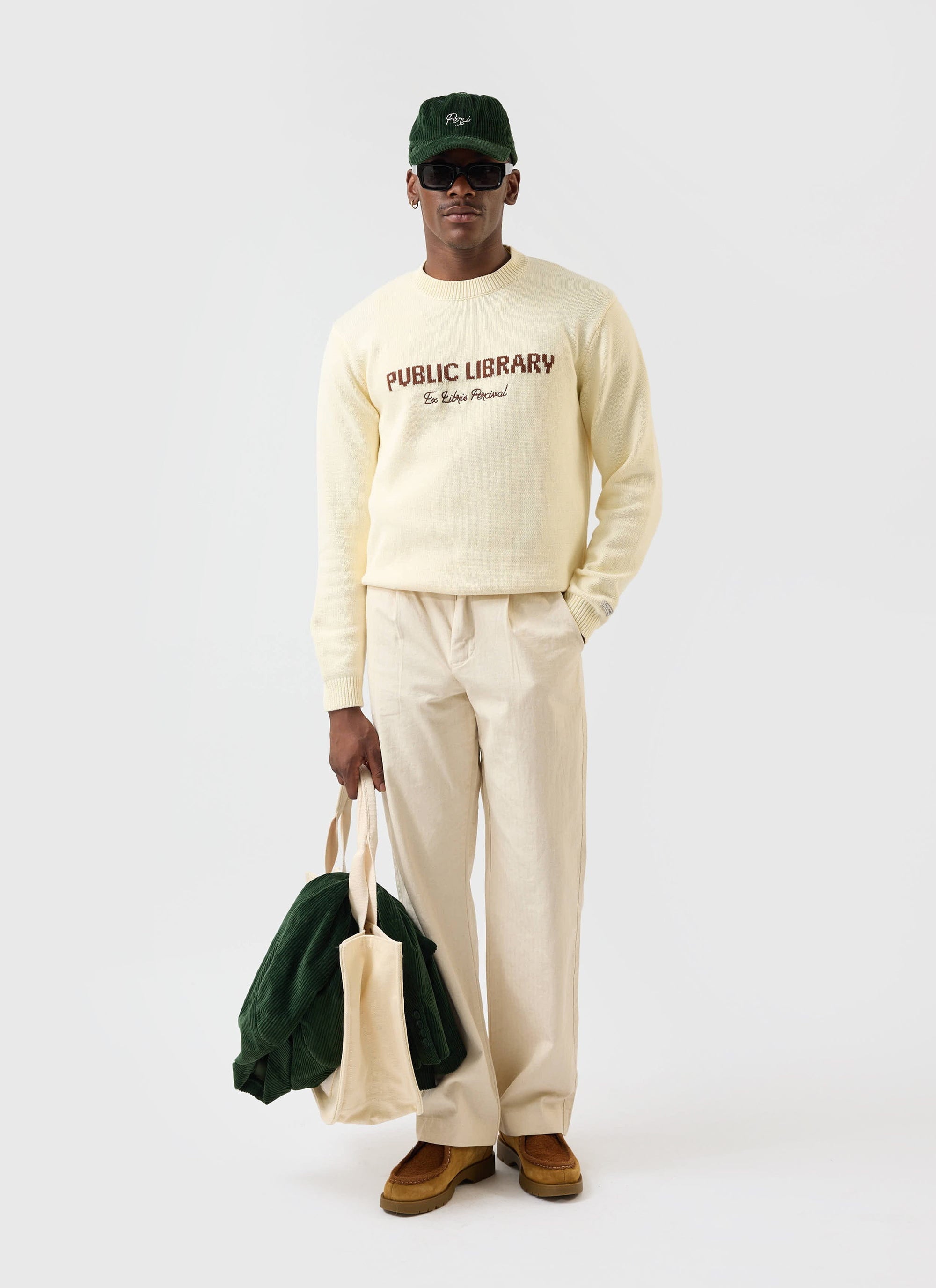 A model wears the ecru cream Public Library knitted jumper from Percival, paired with cream trousers and a green cord cap.