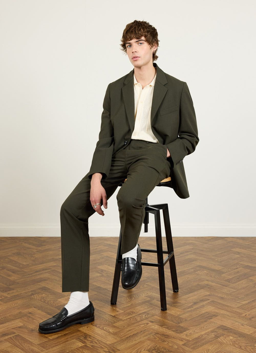 A model sits on a stool wearing the Percival forest green wool suit, styled with a cream knit polo and black loafers.