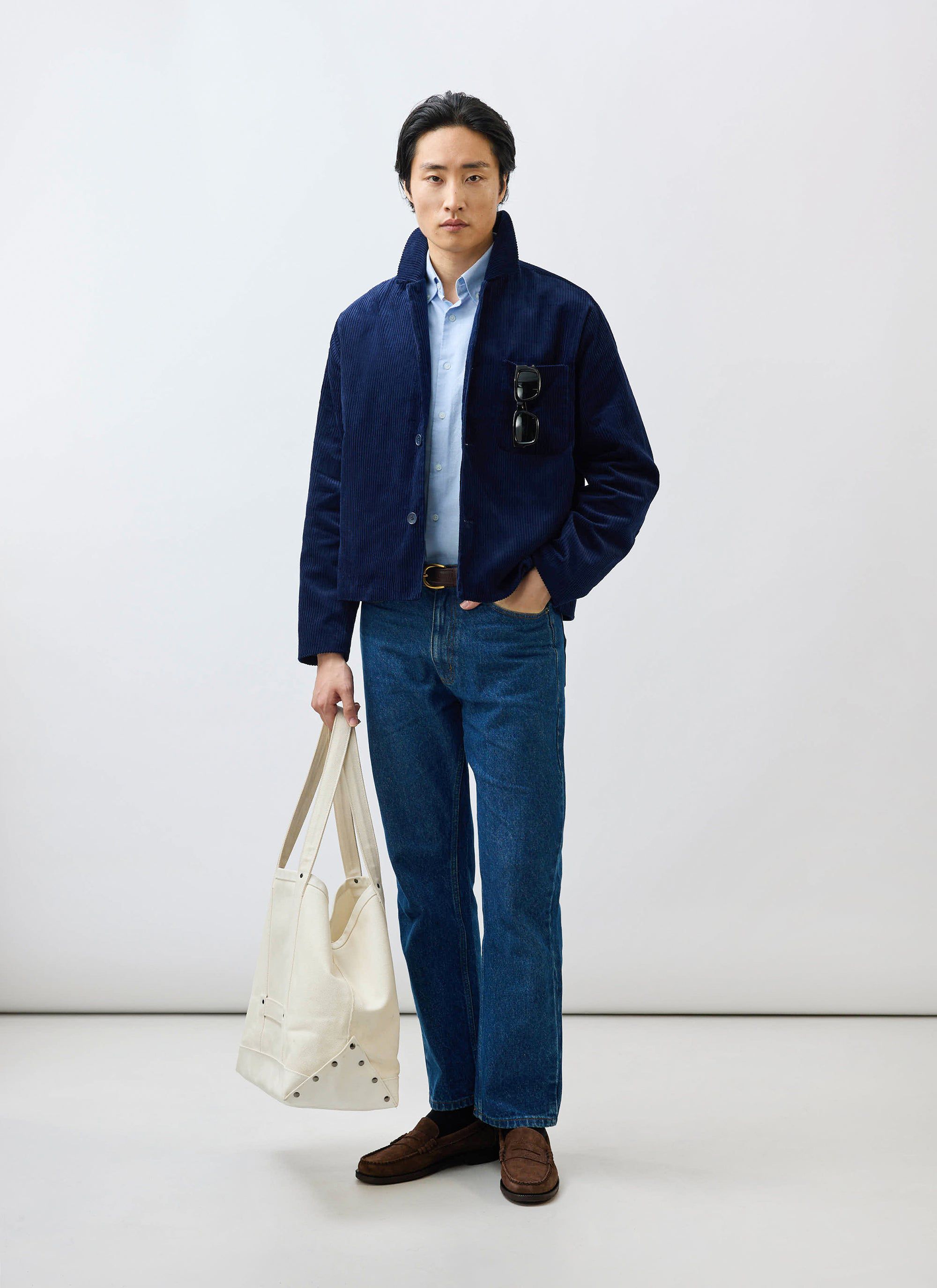 A male model wears the Percival navy corduroy overshirt layered over a blue shirt, with blue jeans and a cream tote bag.