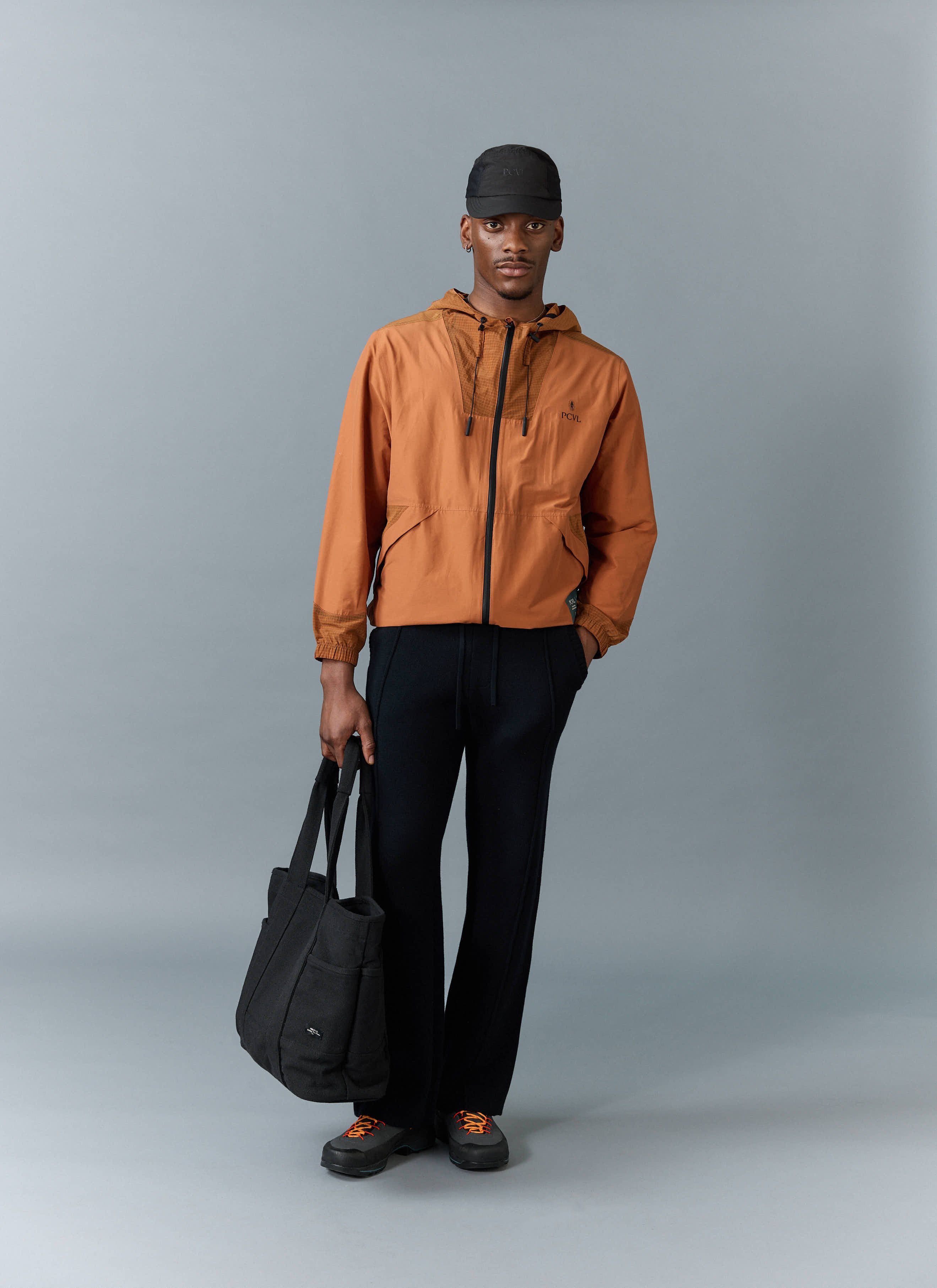A model wears the Percival PCVL Zip Through Water Resistant Jacket in rust, styled with black trousers, a cap and a tote bag.