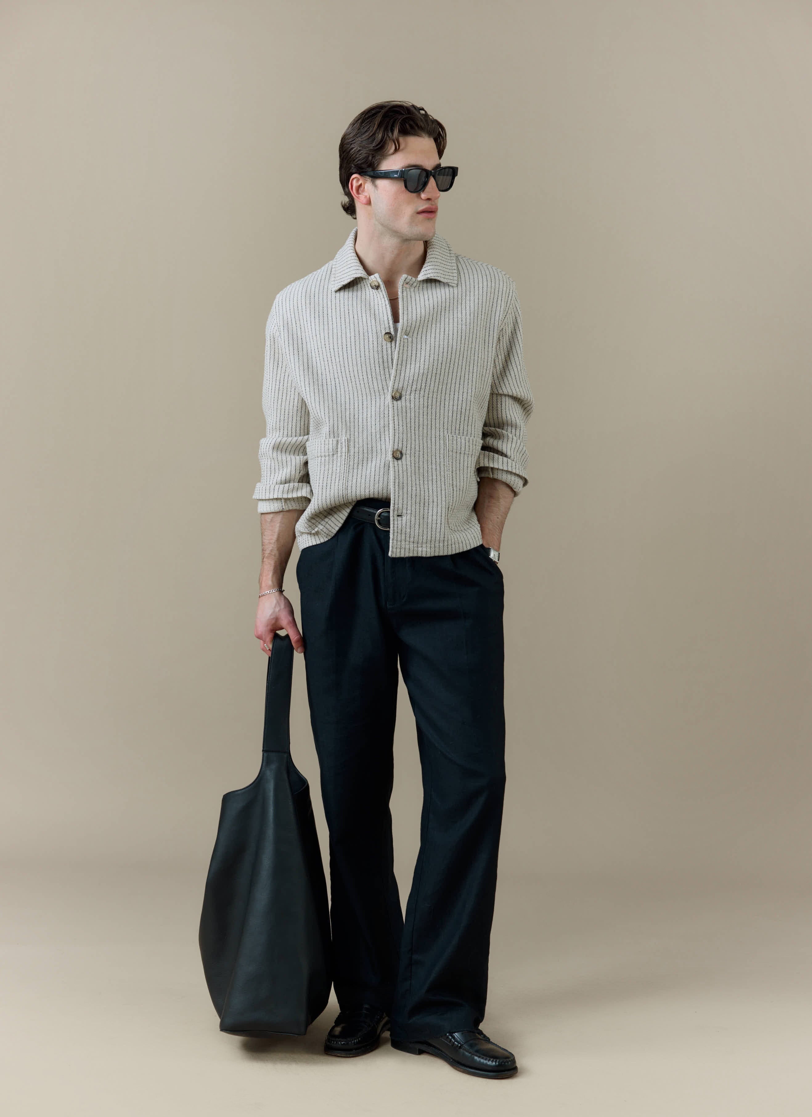 Model wearing the Percival Sorrento Boxy Overshirt in cream linen, paired with black trousers and a large black bag.