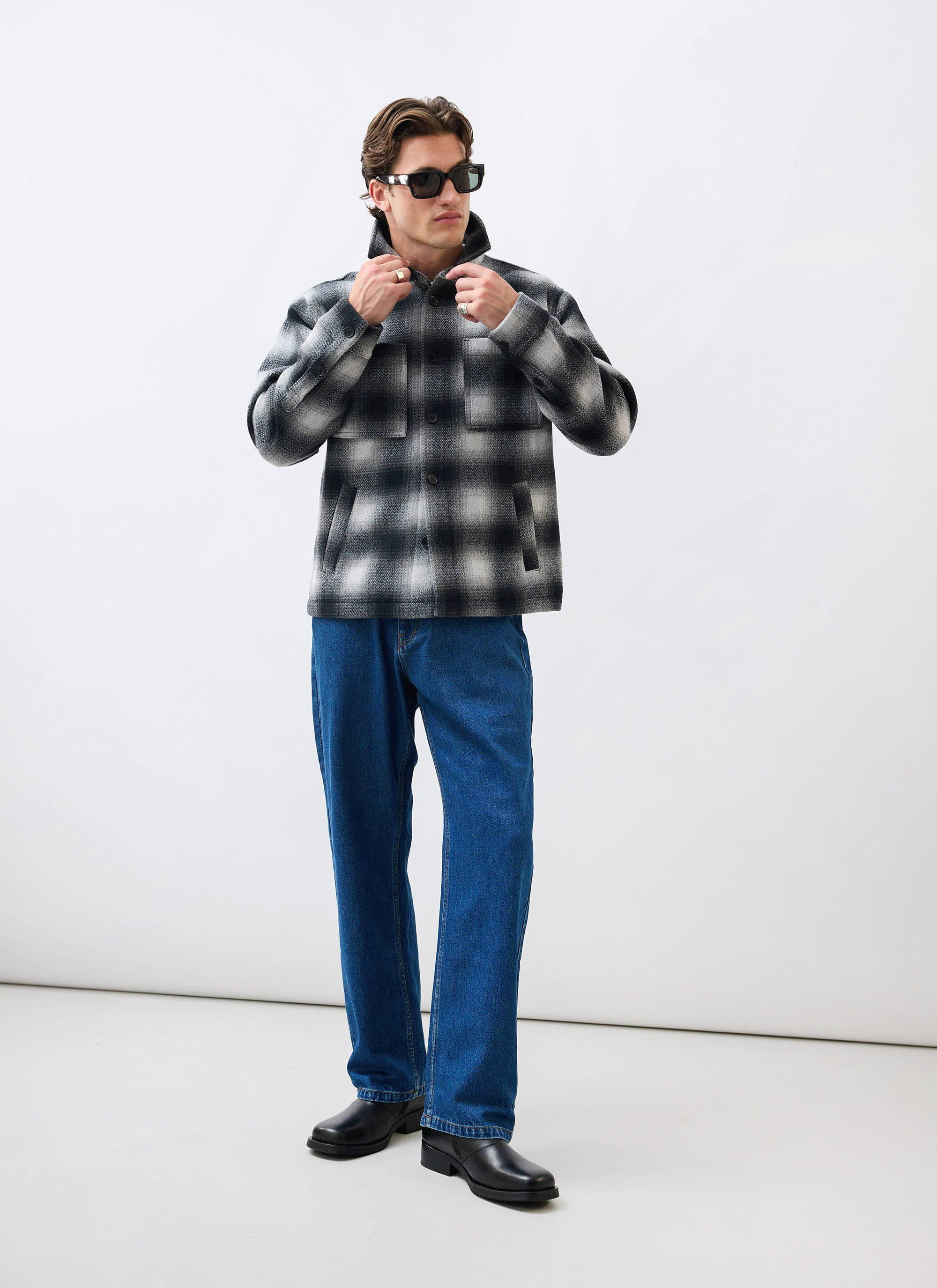A man models the Percival Check Overshirt in grey, styled with dark blue jeans, black shoes, and sunglasses.
