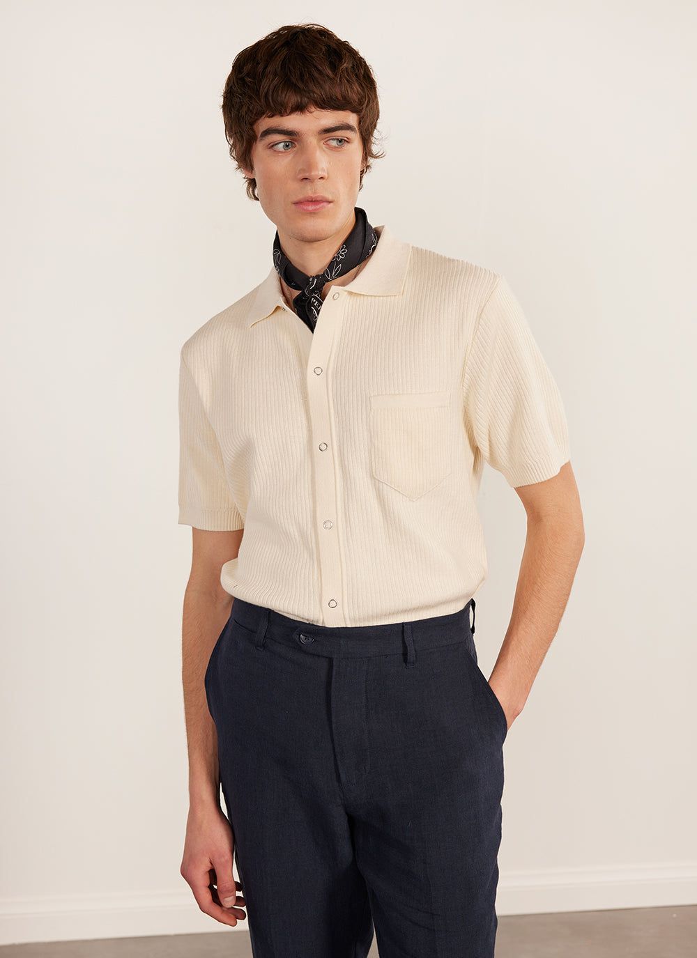 A model wears the Percival Pablo Cuban Shirt in cream, a knitted short sleeve shirt, with dark trousers and a neckerchief.