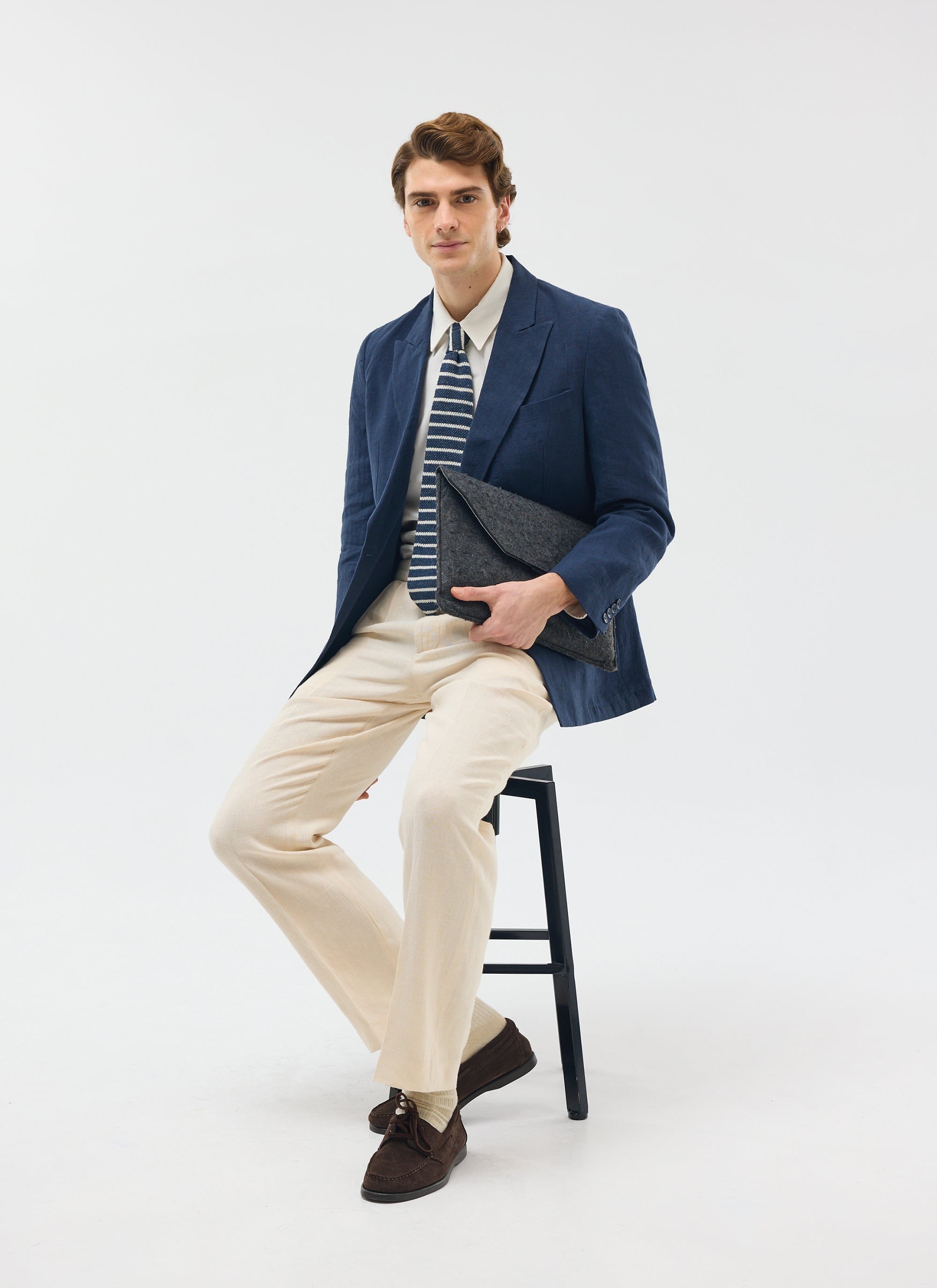 Model wears the Percival navy linen double breasted blazer with cream trousers, a striped tie and holding a grey clutch.
