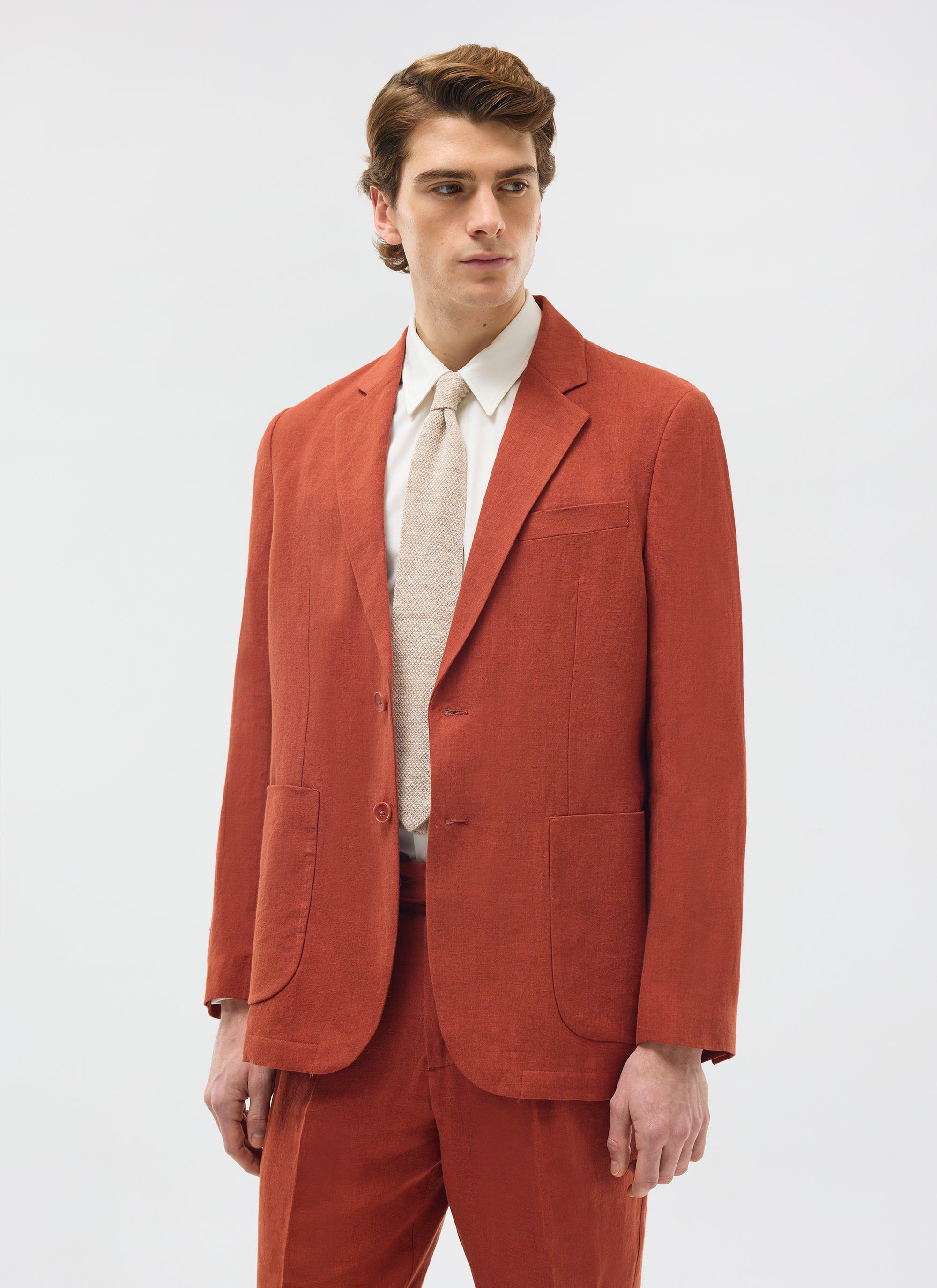 A model wears the Percival Relaxed Tailored Linen Blazer in Rust, with matching trousers, a white shirt and a tie.