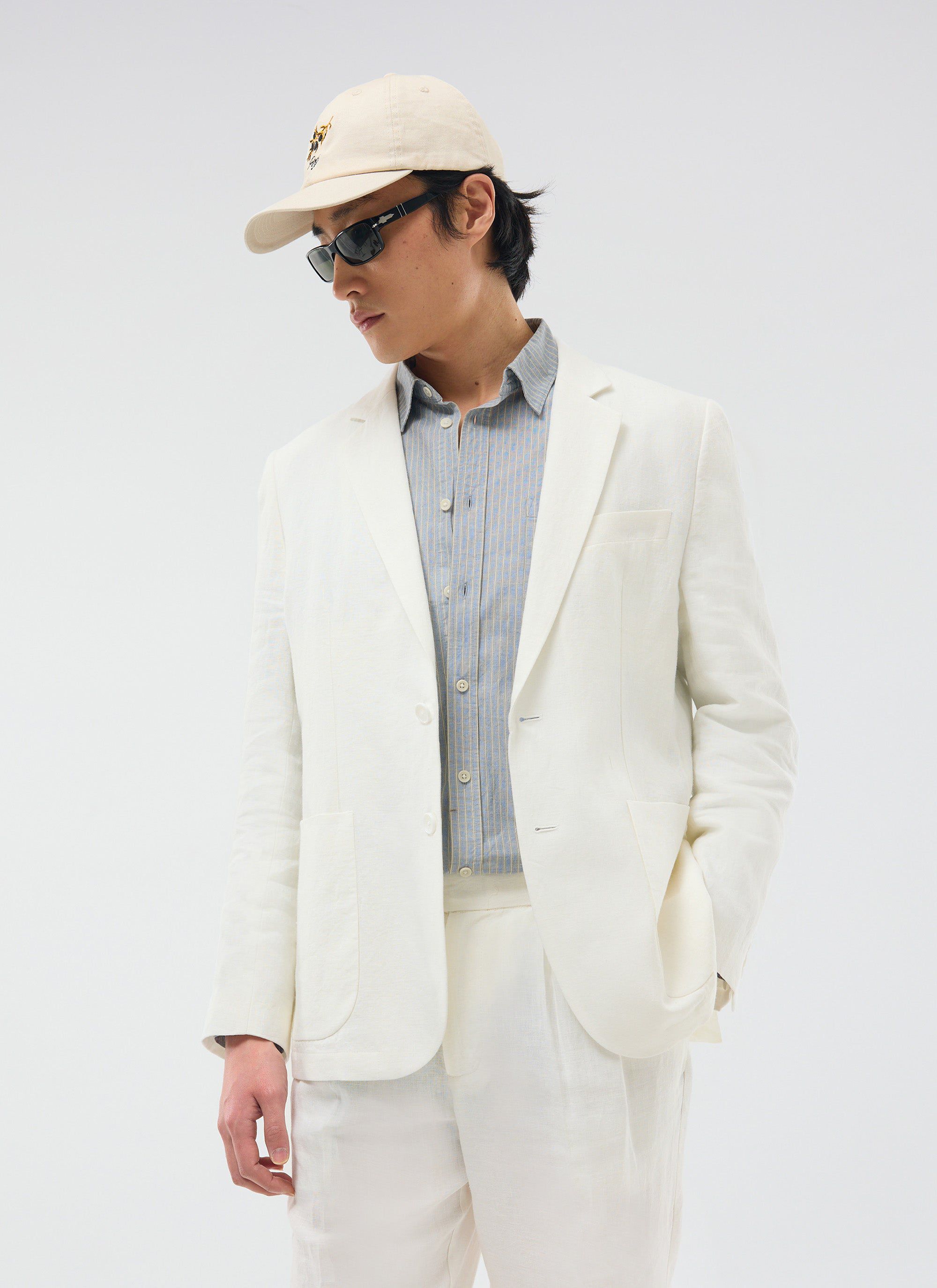 A model wears the Percival Relaxed Tailored Linen Blazer in white, styled casually with a striped shirt and a baseball cap.