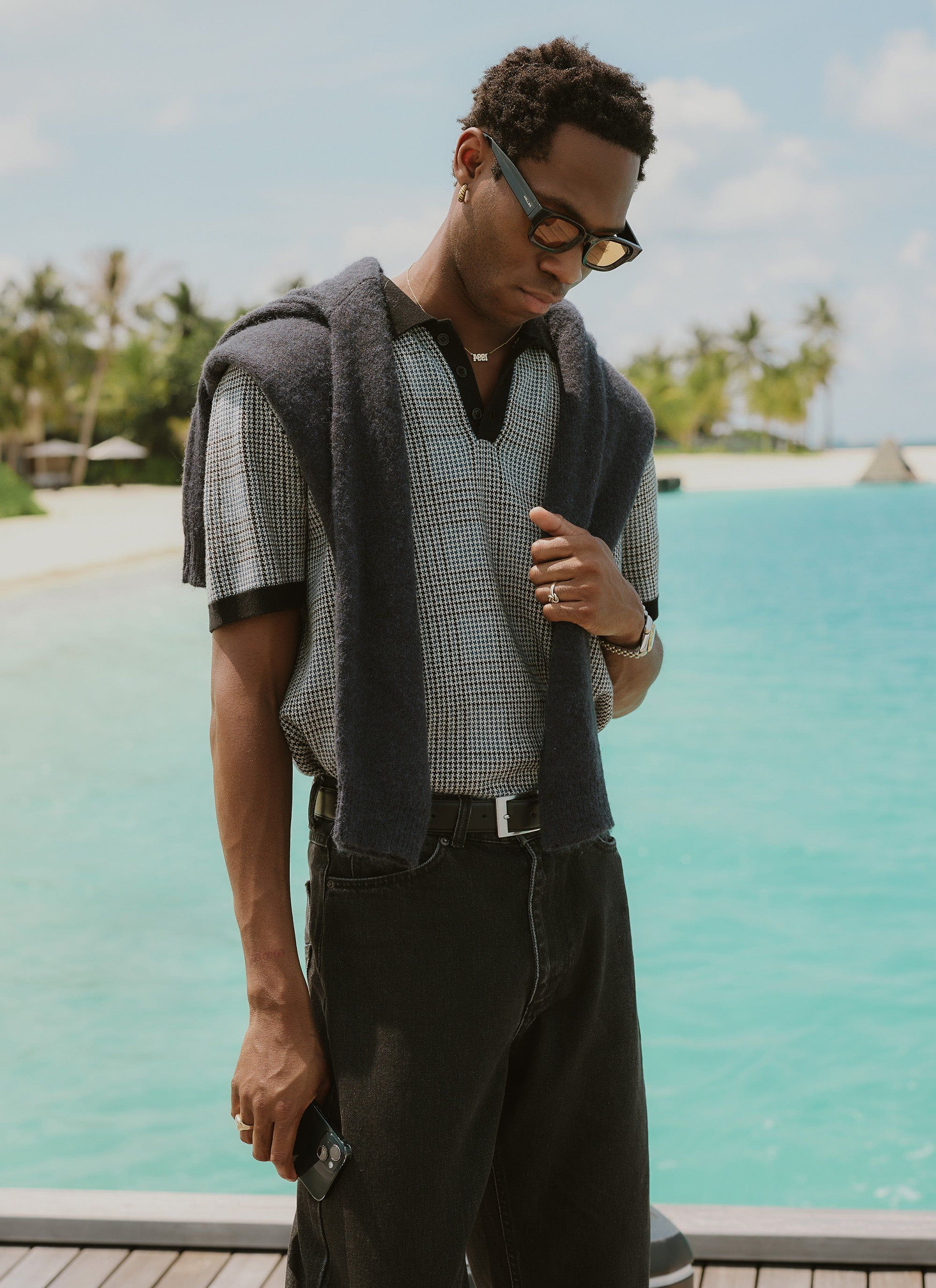 A man wearing the Percival Casa Martini Knitted Polo in a navy puppytooth pattern, standing on a deck overlooking the sea.